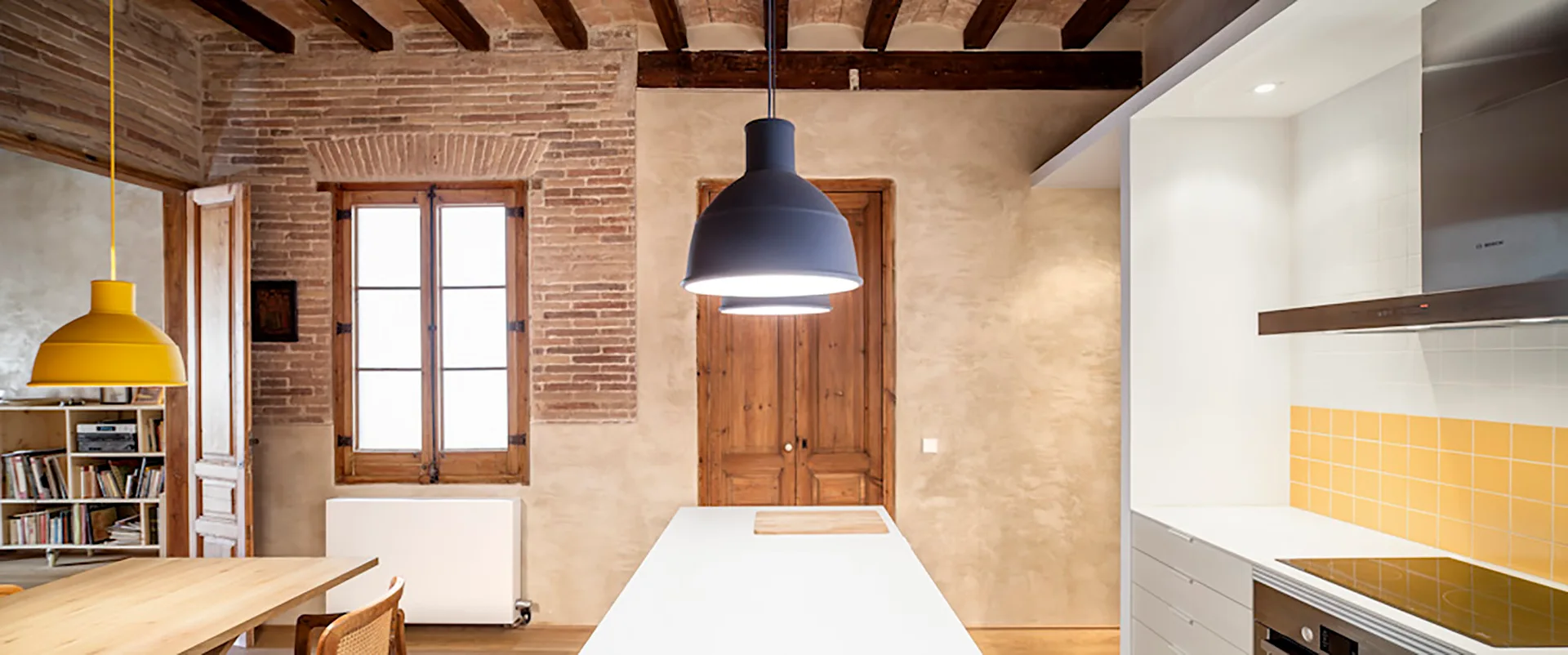 A modern kitchen featuring a minimalist white island, yellow tiled backsplash, exposed brick walls, and rustic wooden ceiling beams.