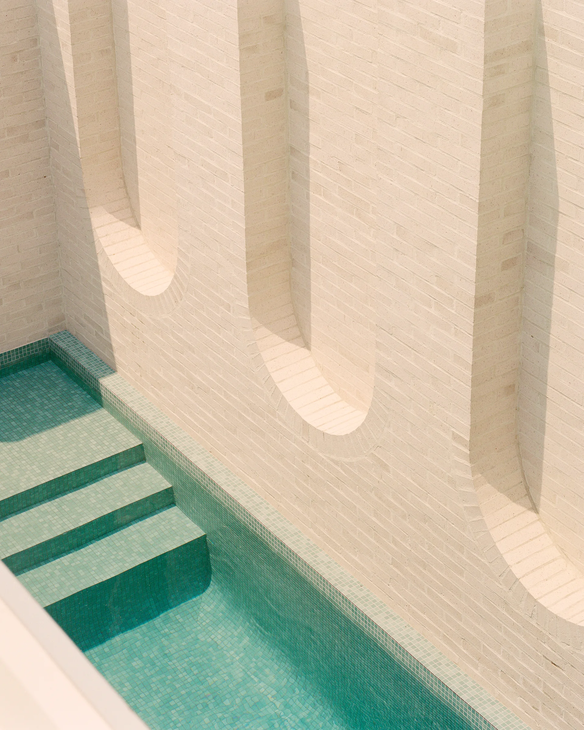 A high-angle view of a turquoise mosaic tiled pool with steps next to a cream brick wall featuring deep arched architectural recesses.
