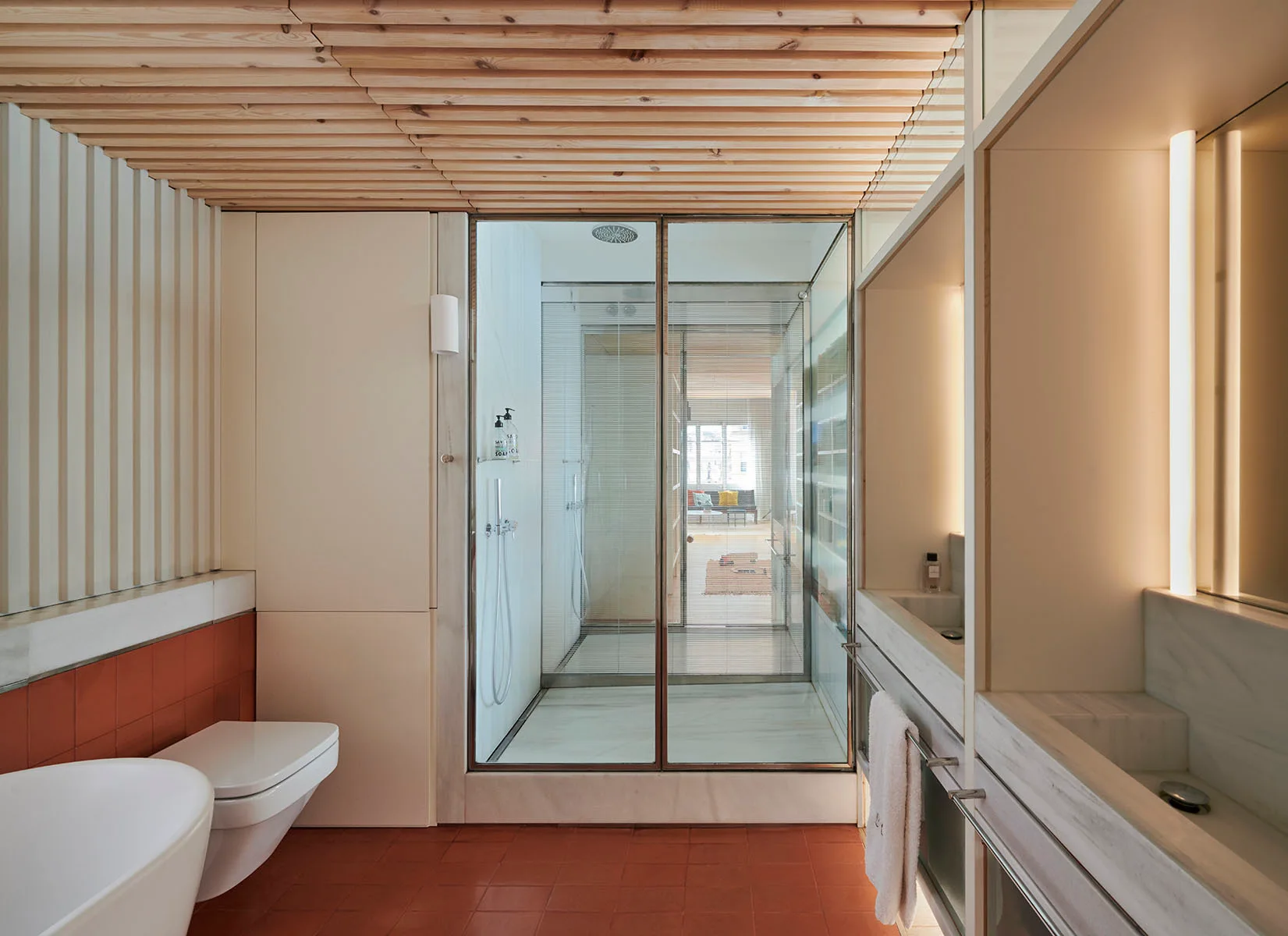 A contemporary bathroom features a slatted wood ceiling, terracotta tile floor, a glass-enclosed shower, and a marble vanity.