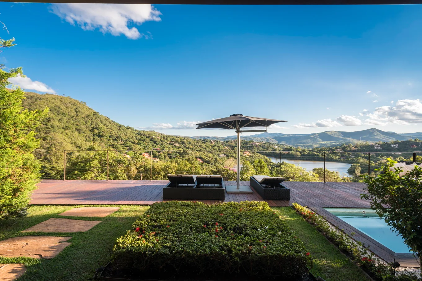 A spacious wooden deck terrace features dark wicker sun loungers under a black patio umbrella overlooking a scenic mountain lake landscape.