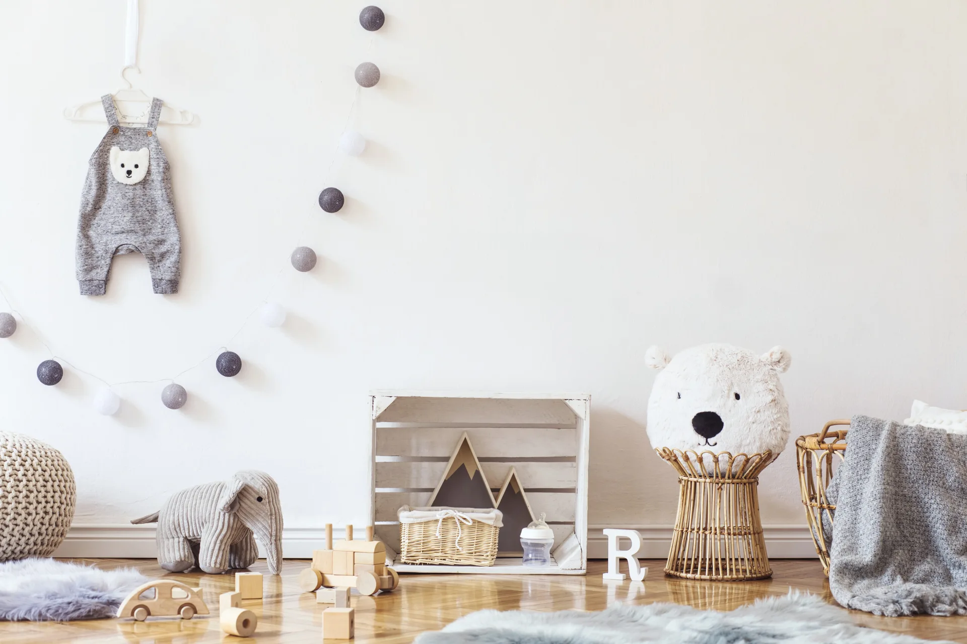 A minimalist nursery features wooden toys, a storage crate, a wicker stool with a bear pillow, and a baby jumpsuit on a white wall.