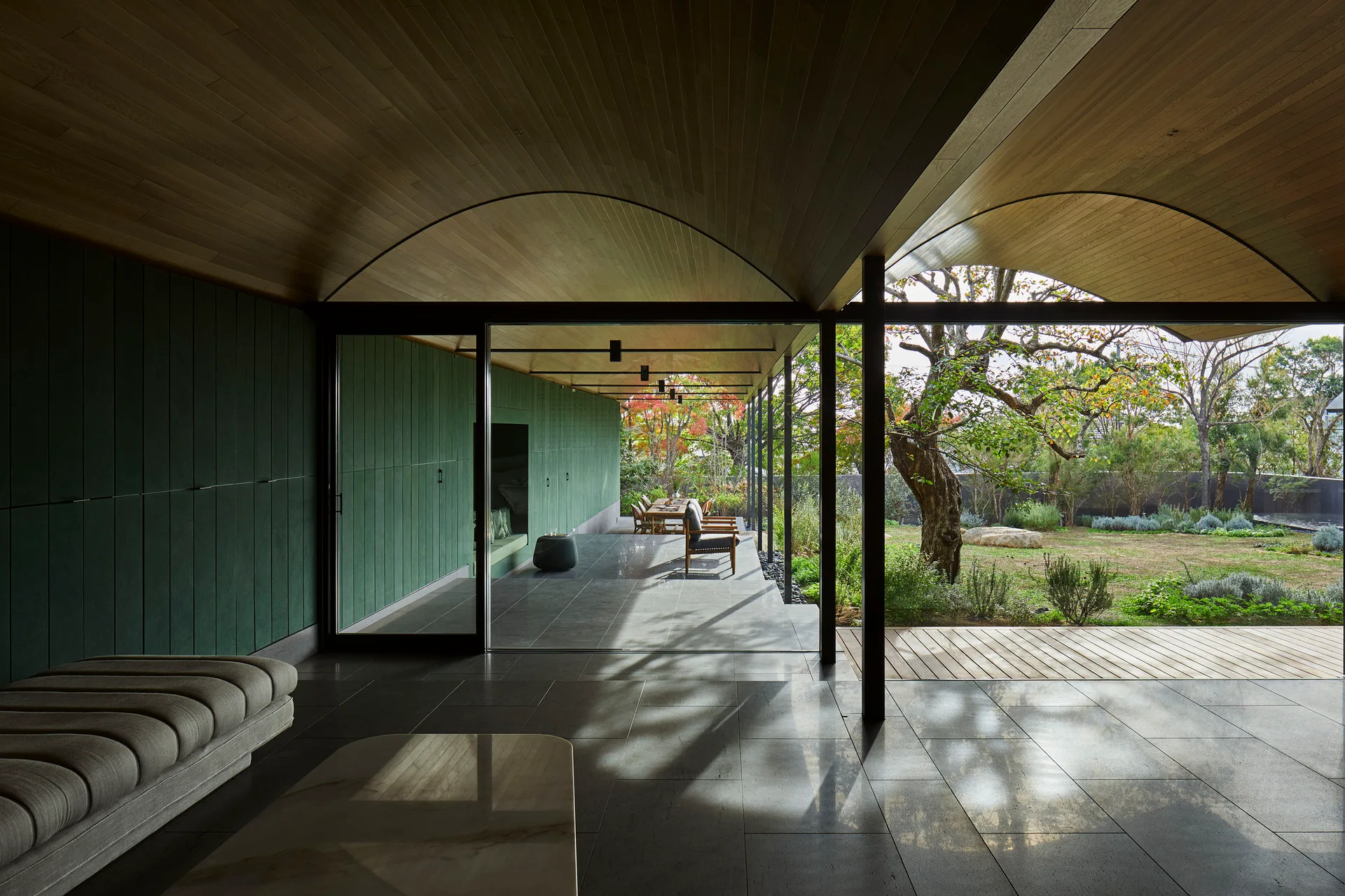A modern living room with a barrel vaulted wood ceiling, green paneled wall, and glass sliding doors that open to a stone tile terrace and garden.