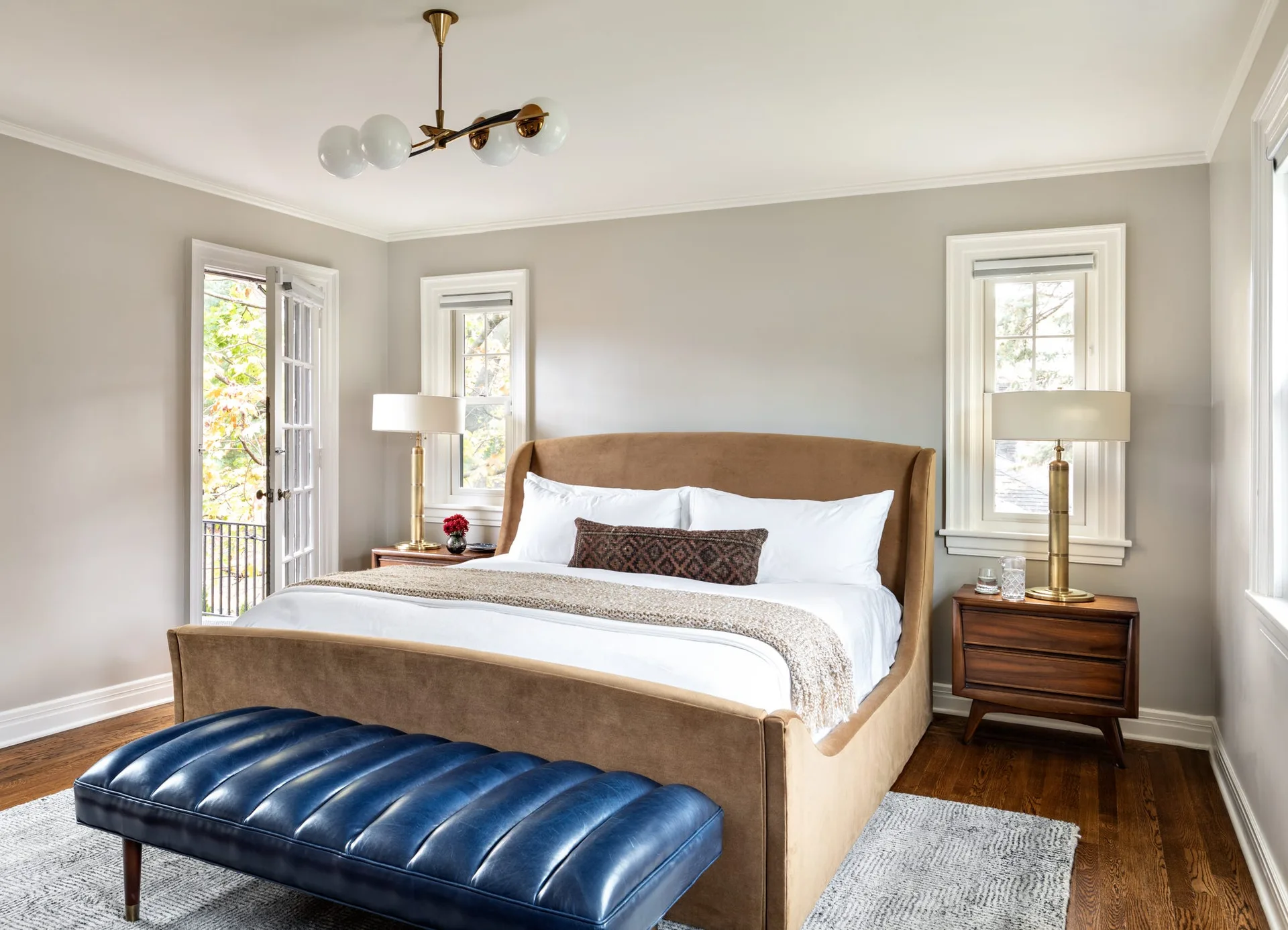 A bedroom with light grey walls features a brown upholstered bed, mid-century wooden nightstands with brass lamps, and a blue tufted bench at the foot.
