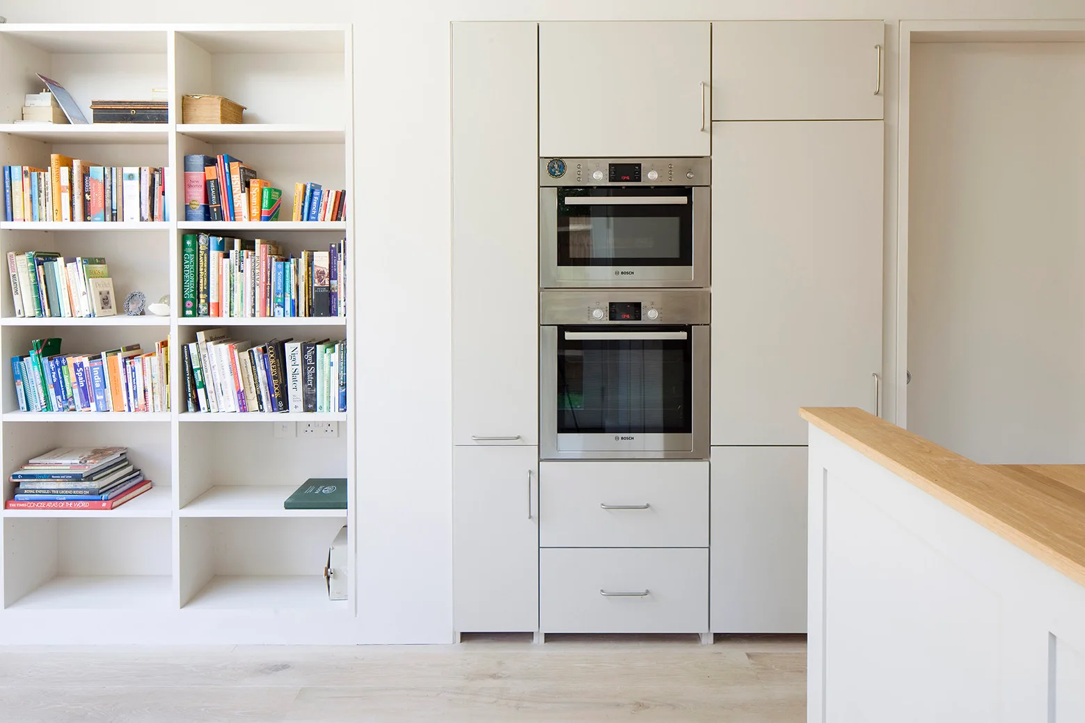 A modern white kitchen featuring an integrated double wall oven next to a built-in bookshelf filled with books and a light wood countertop.