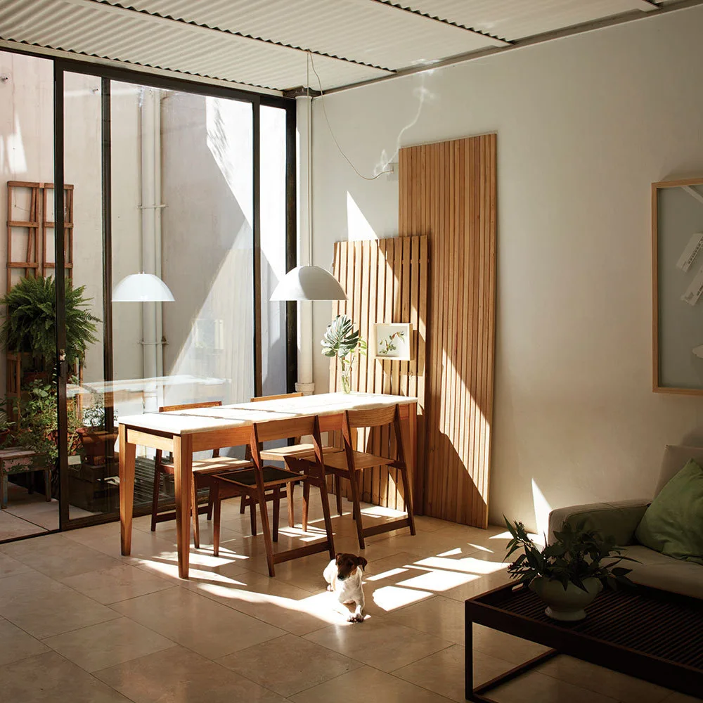 A sunlit dining area with a wooden table, chairs, leaning slatted wood screens against a white wall, and a large sliding glass door opening to a patio.
