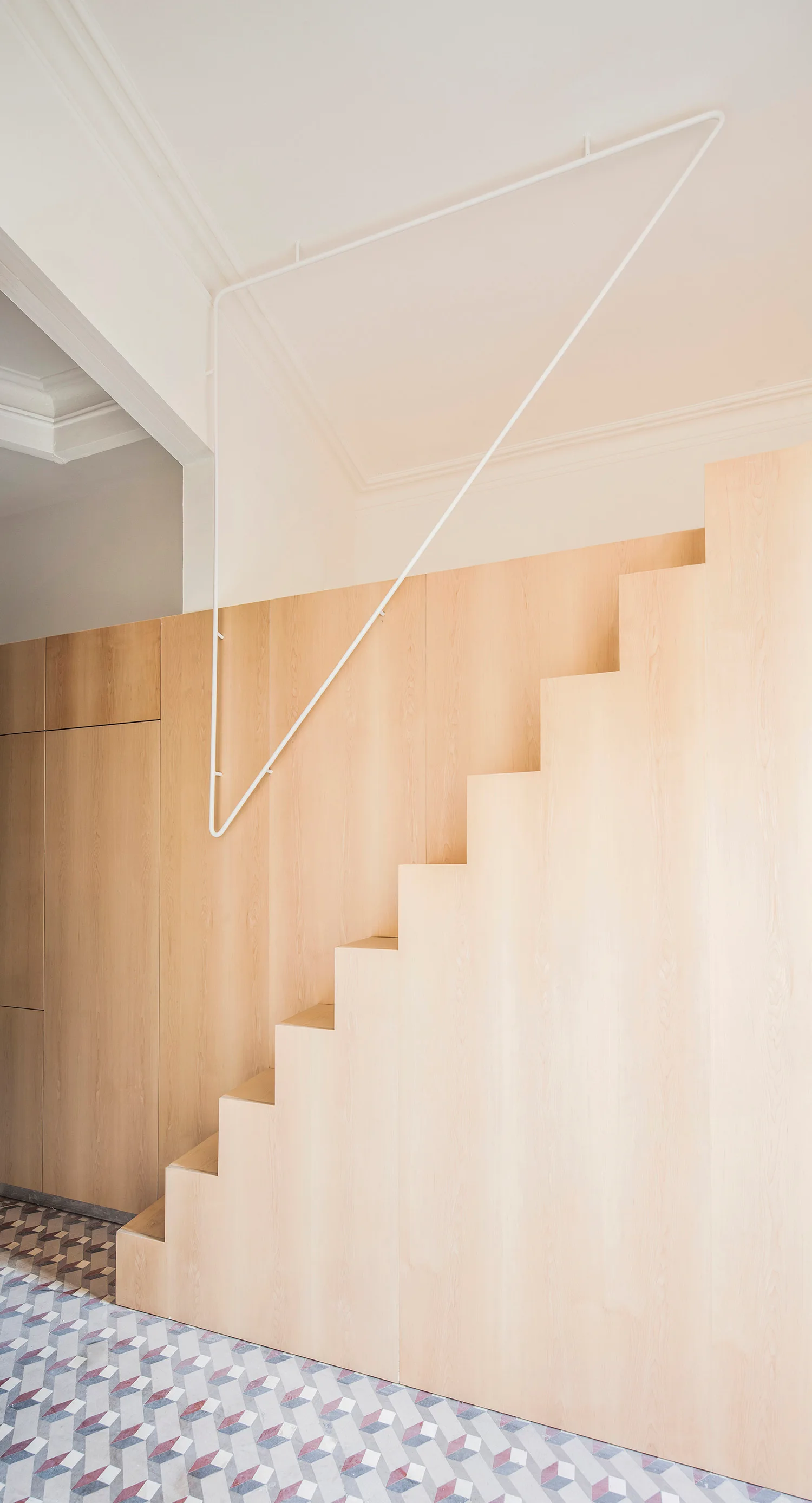 A minimalist light wood staircase featuring integrated storage cabinets stands on a geometric patterned tile floor beneath a white metal handrail.