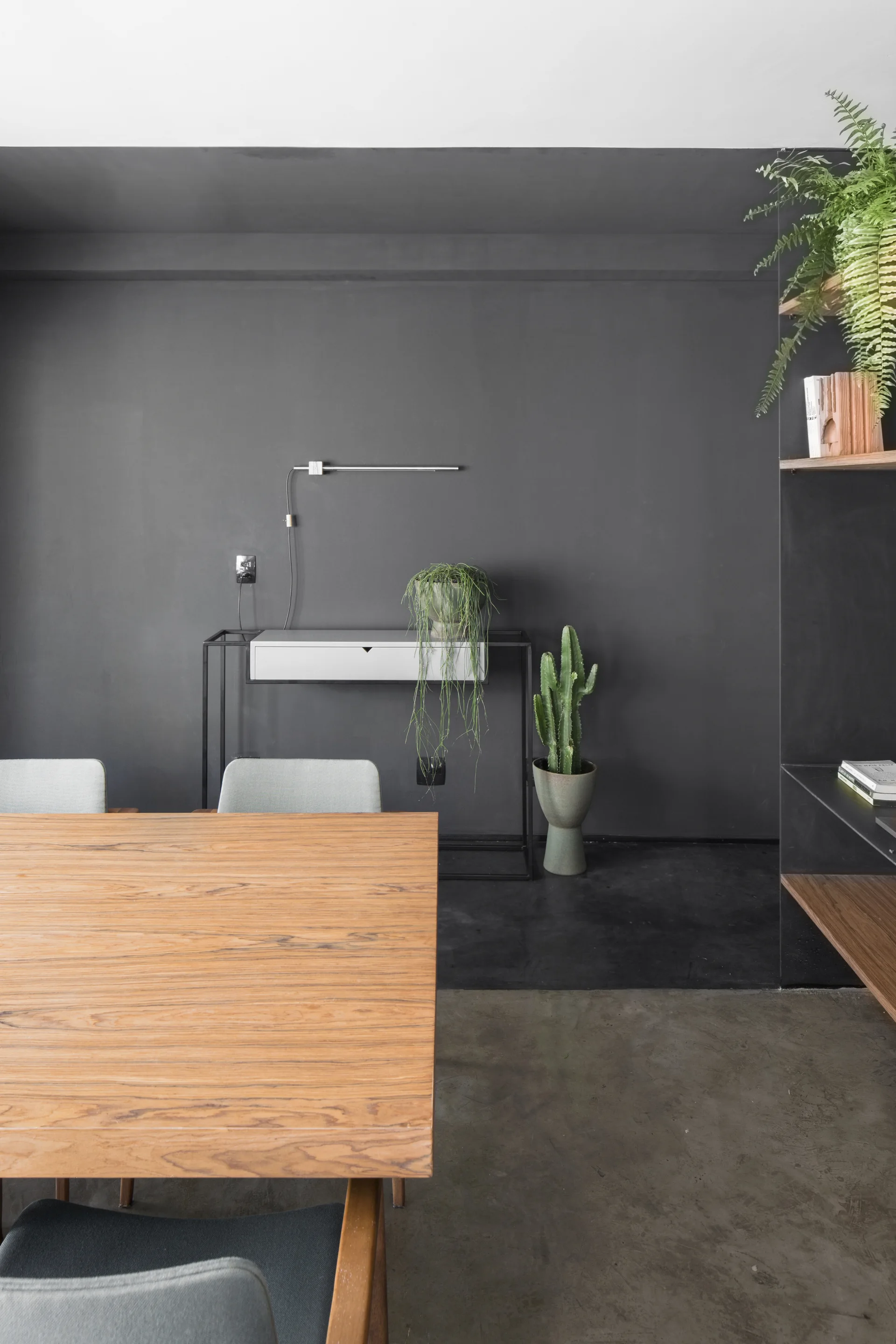 A minimalist dining room features a warm wood table, grey chairs, and a dark grey accent wall with a narrow console and potted plants.