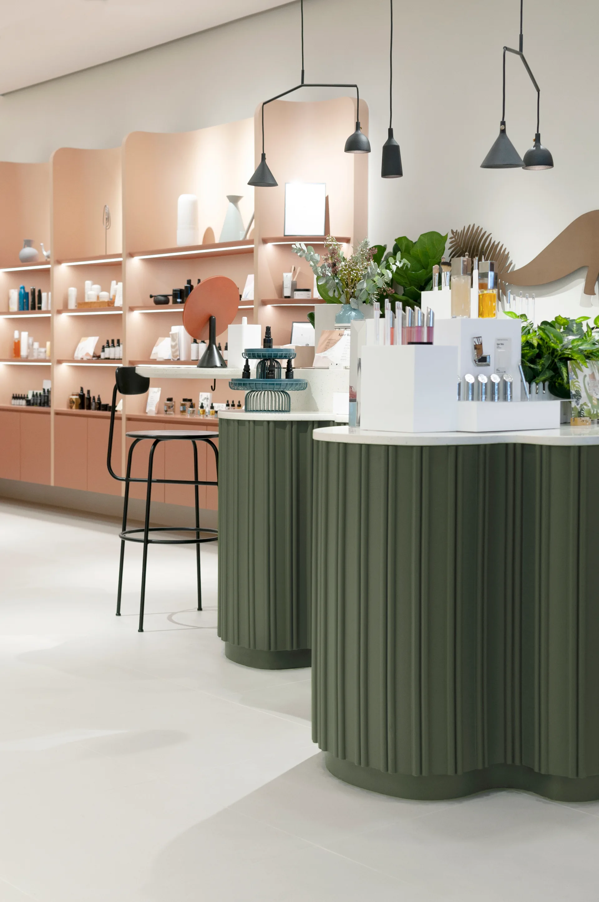A modern interior showing curved olive green fluted counters with light stone tops alongside curved peach shelving units and black pendant lights.