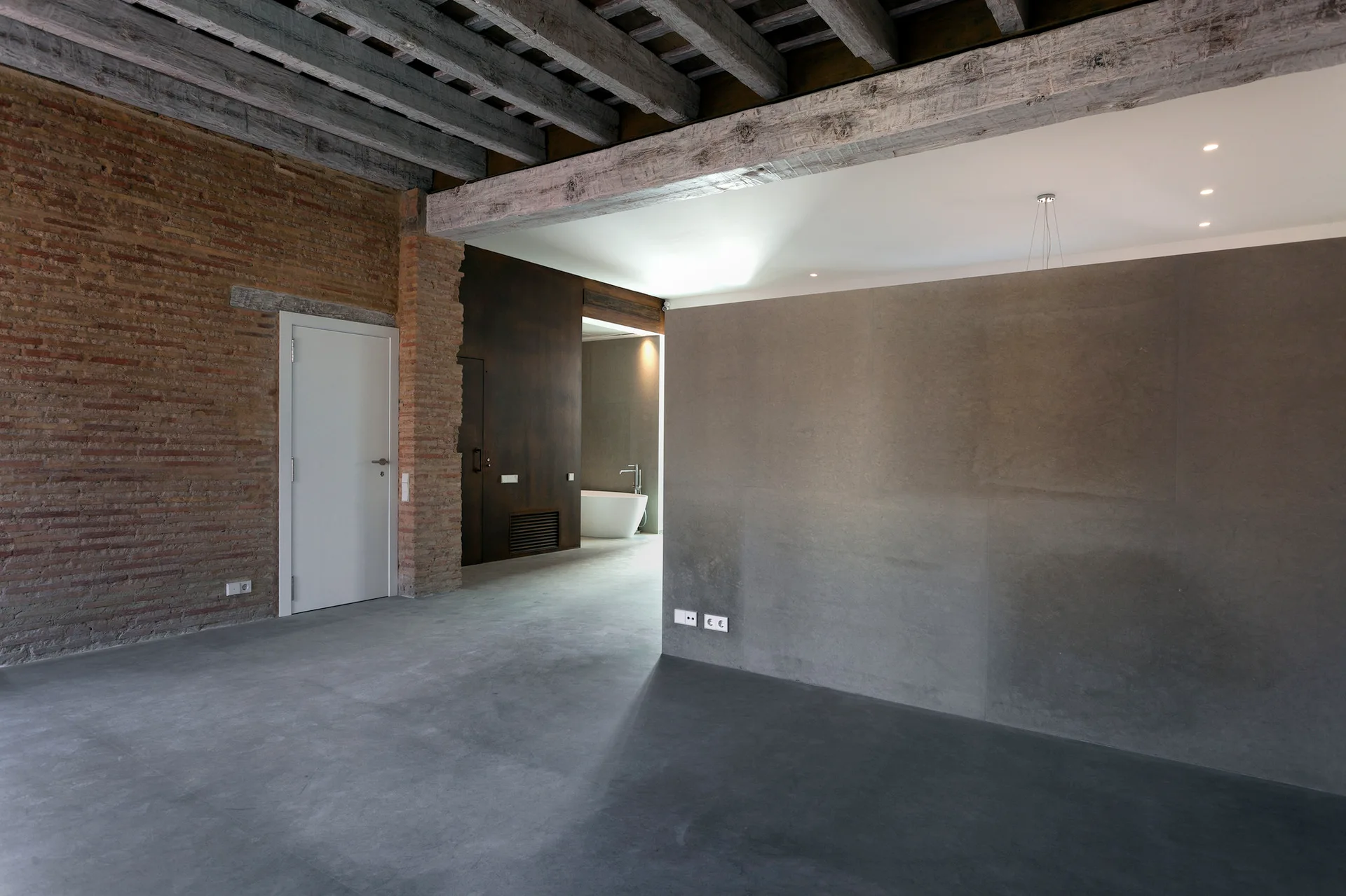 An industrial interior features a smooth concrete floor and partition wall alongside an exposed brick wall, timber ceiling beams, and a white door.