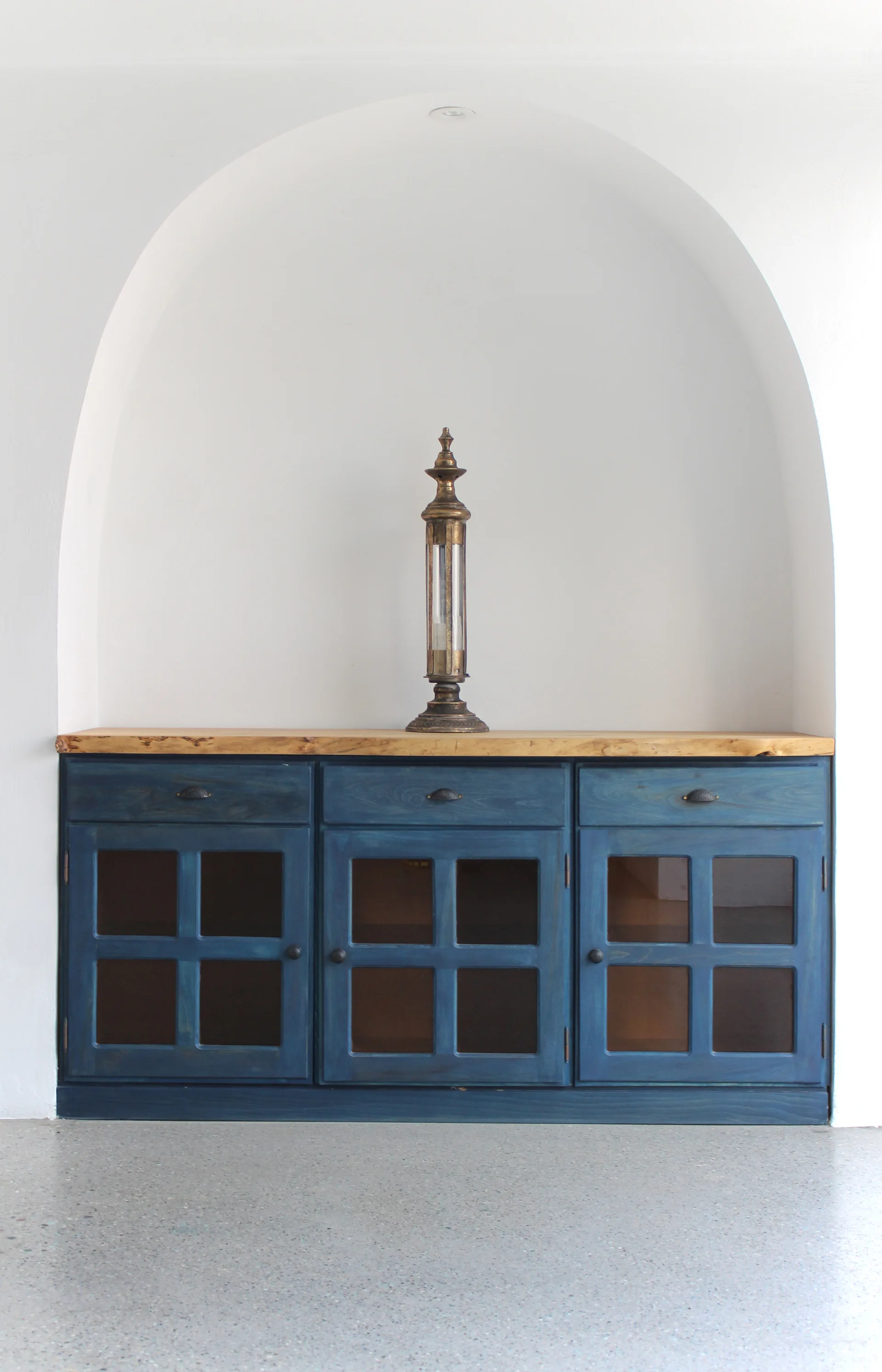 A deep blue wooden cabinet with glass doors and a wood countertop sits inside a white arched wall alcove.
