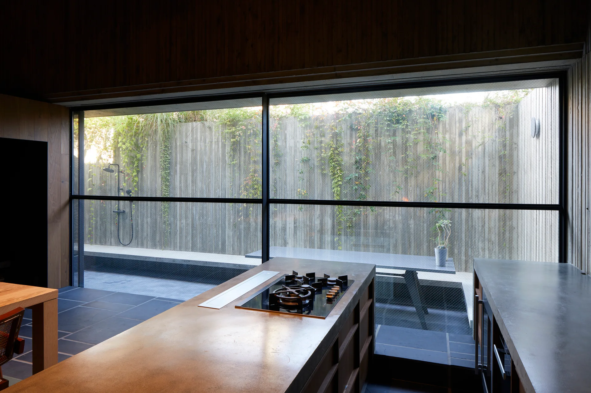 A modern kitchen with a stone-top island and gas cooktop looks out through a large wire-mesh window to a private, vine-covered courtyard.