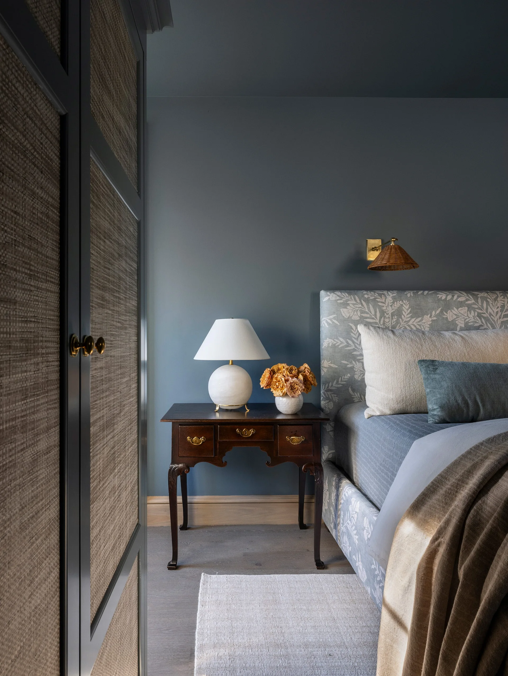 A blue bedroom features a floral upholstered headboard, an antique wood nightstand with a white table lamp, and a textured closet door.