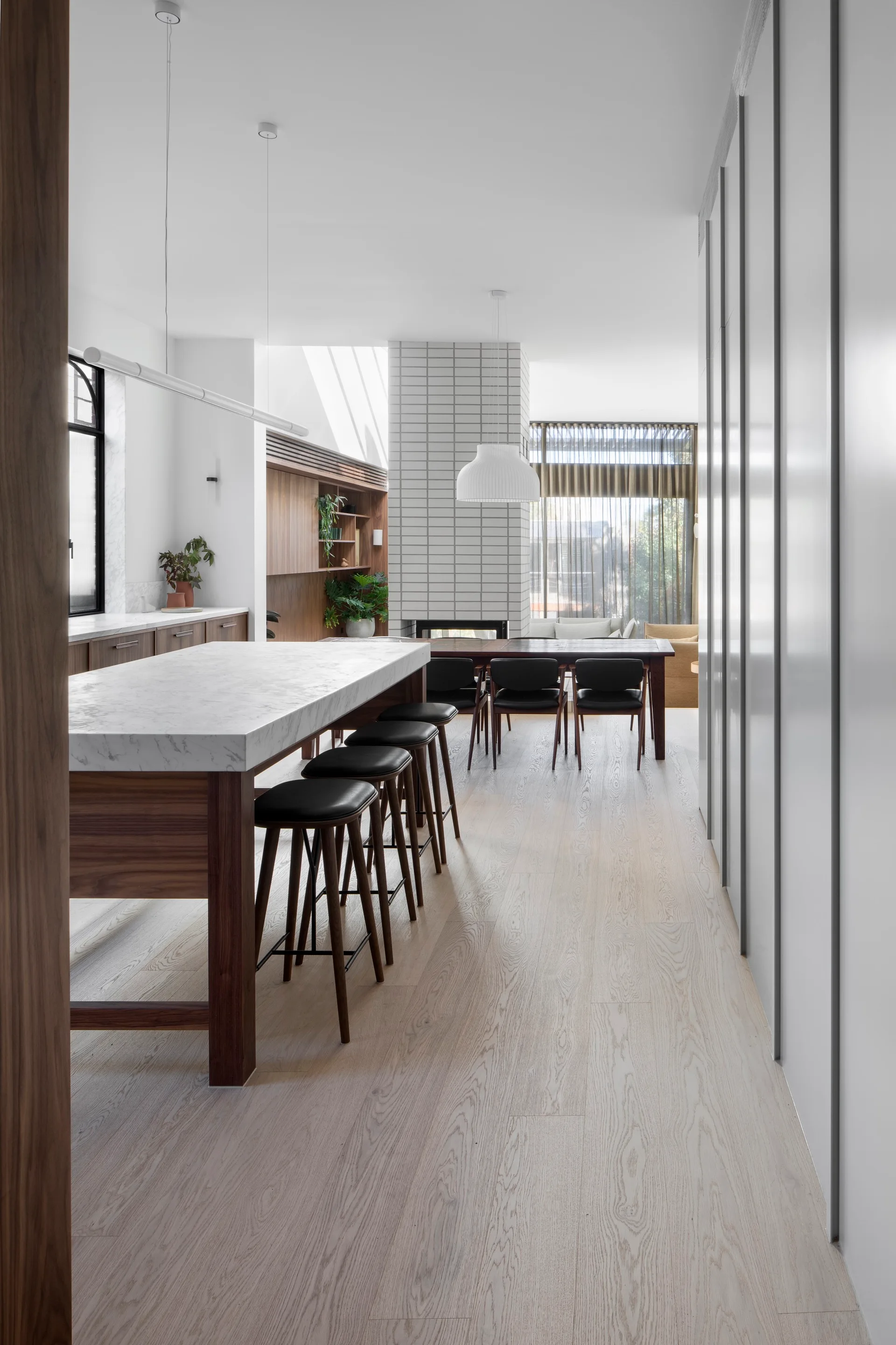 A modern open kitchen features a marble-topped wood island with bar stools and a central white-tiled fireplace column dividing the space.