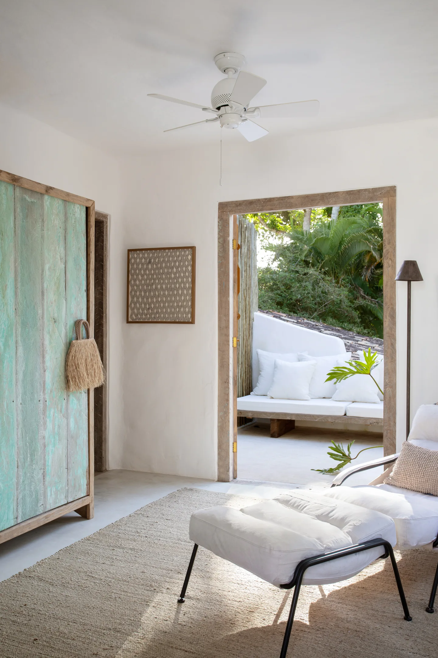 A sunlit bedroom corner features a white lounge chair with ottoman, a woven jute rug, and a distressed green wooden wardrobe next to an open doorway.