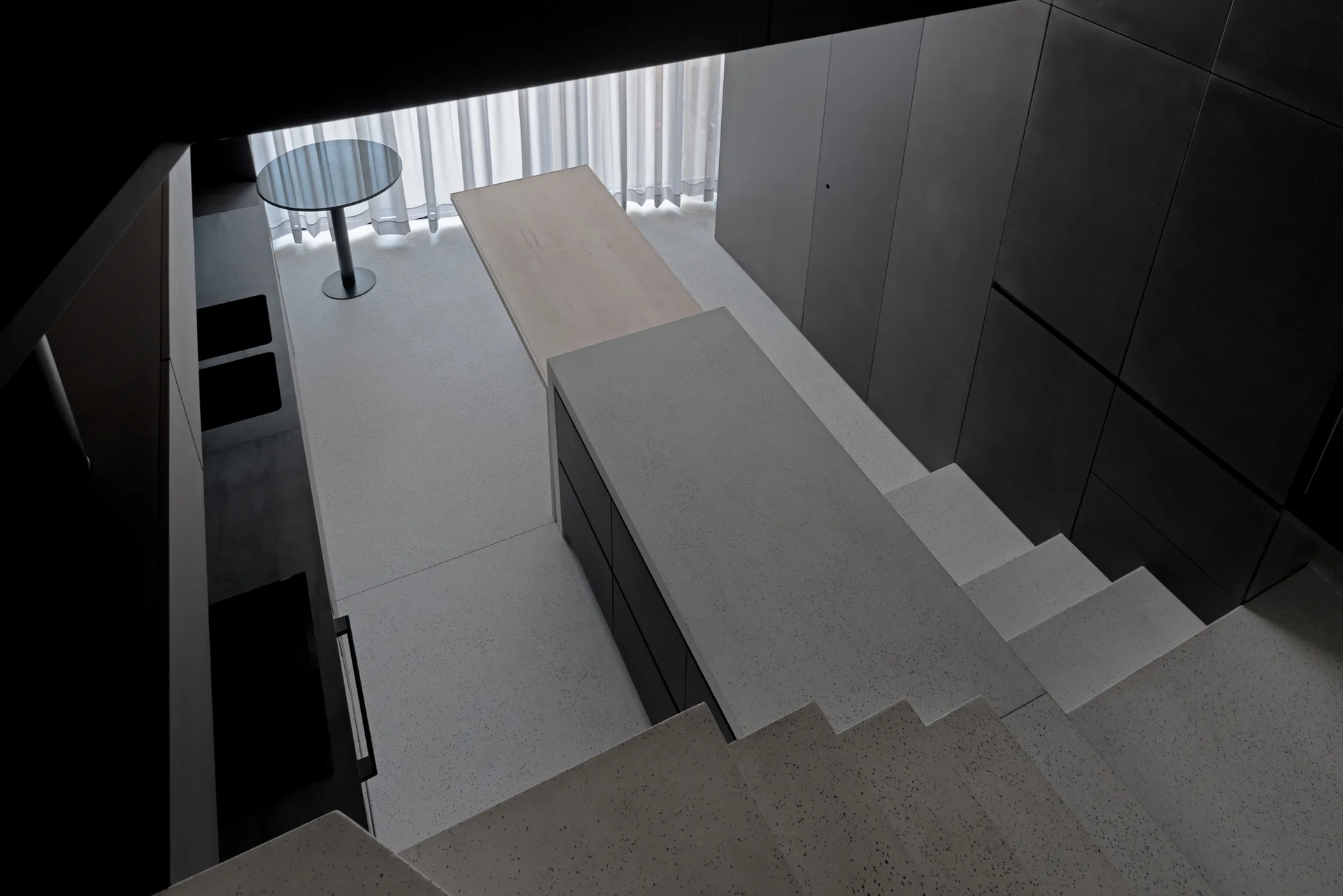High-angle view of a minimalist kitchen showing a terrazzo stone staircase leading down to a dark grey island with a wood extension.