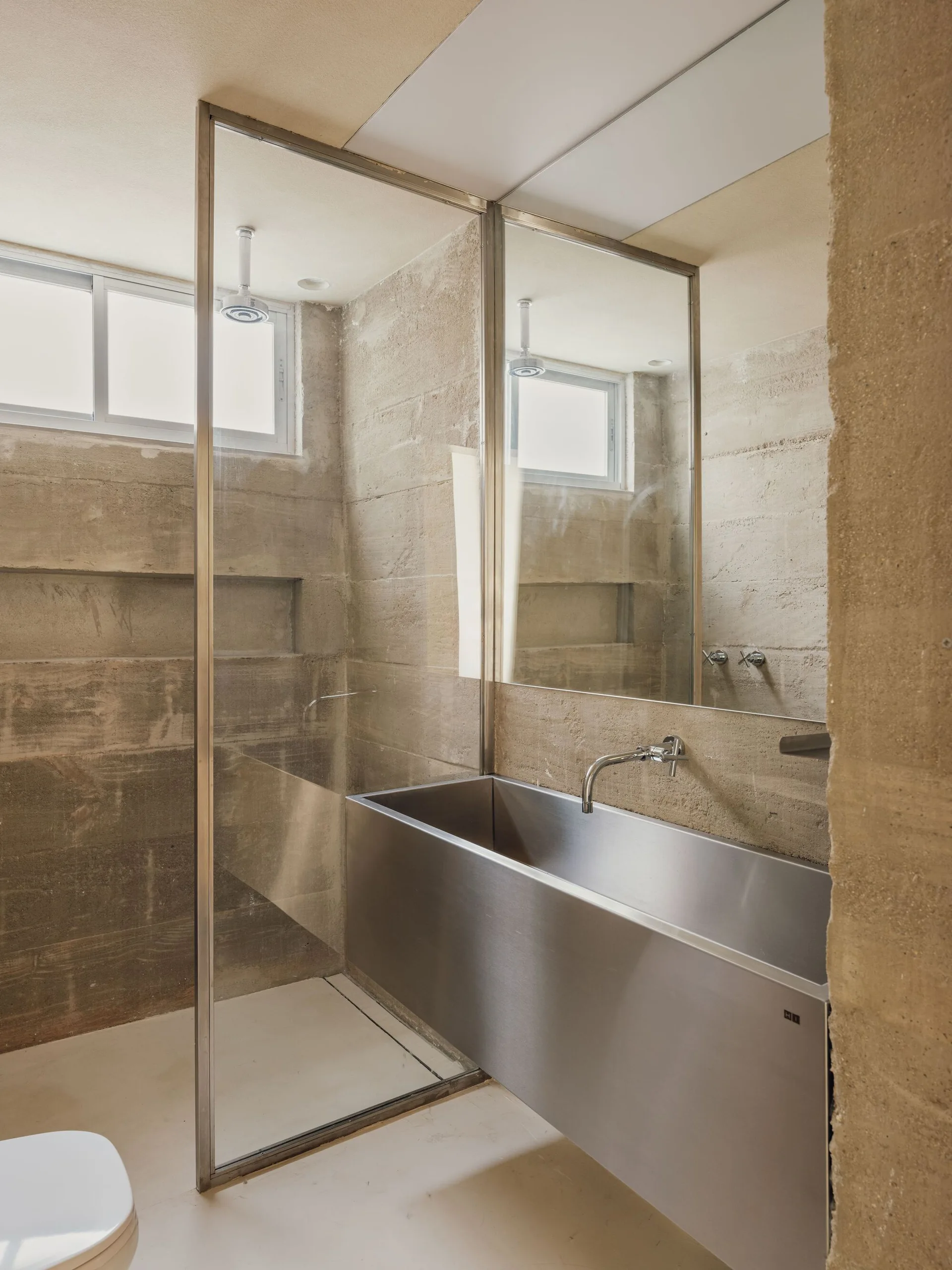 A minimalist bathroom featuring raw beige concrete walls, a wall-mounted stainless steel trough sink, and a clear glass shower screen.