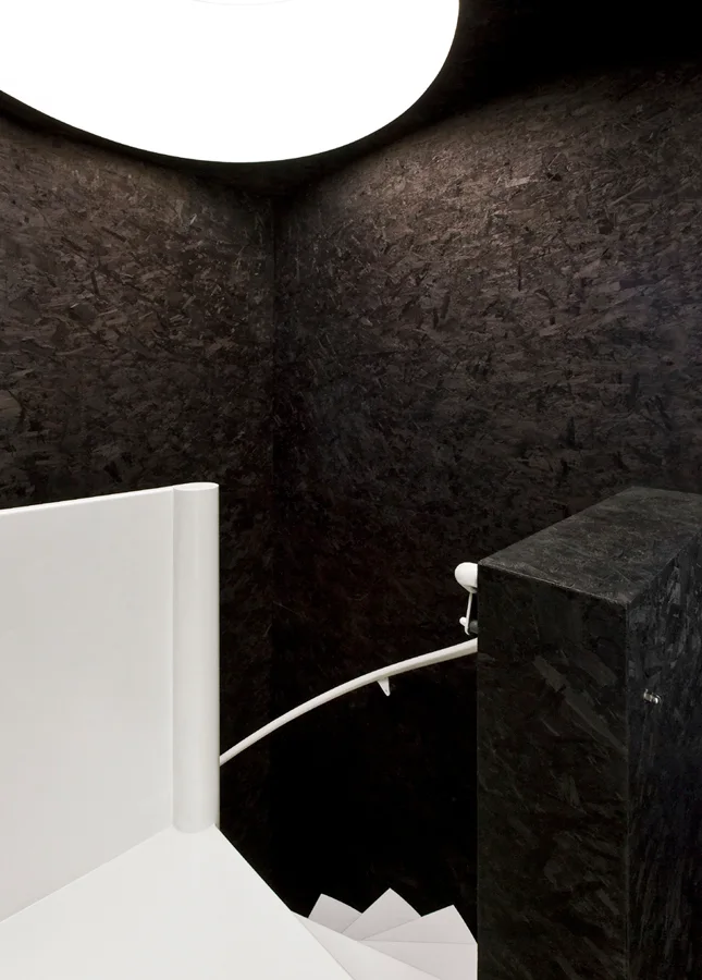 A stark stairwell features matte black oriented strand board walls, a winding white staircase, and a matching curved handrail.