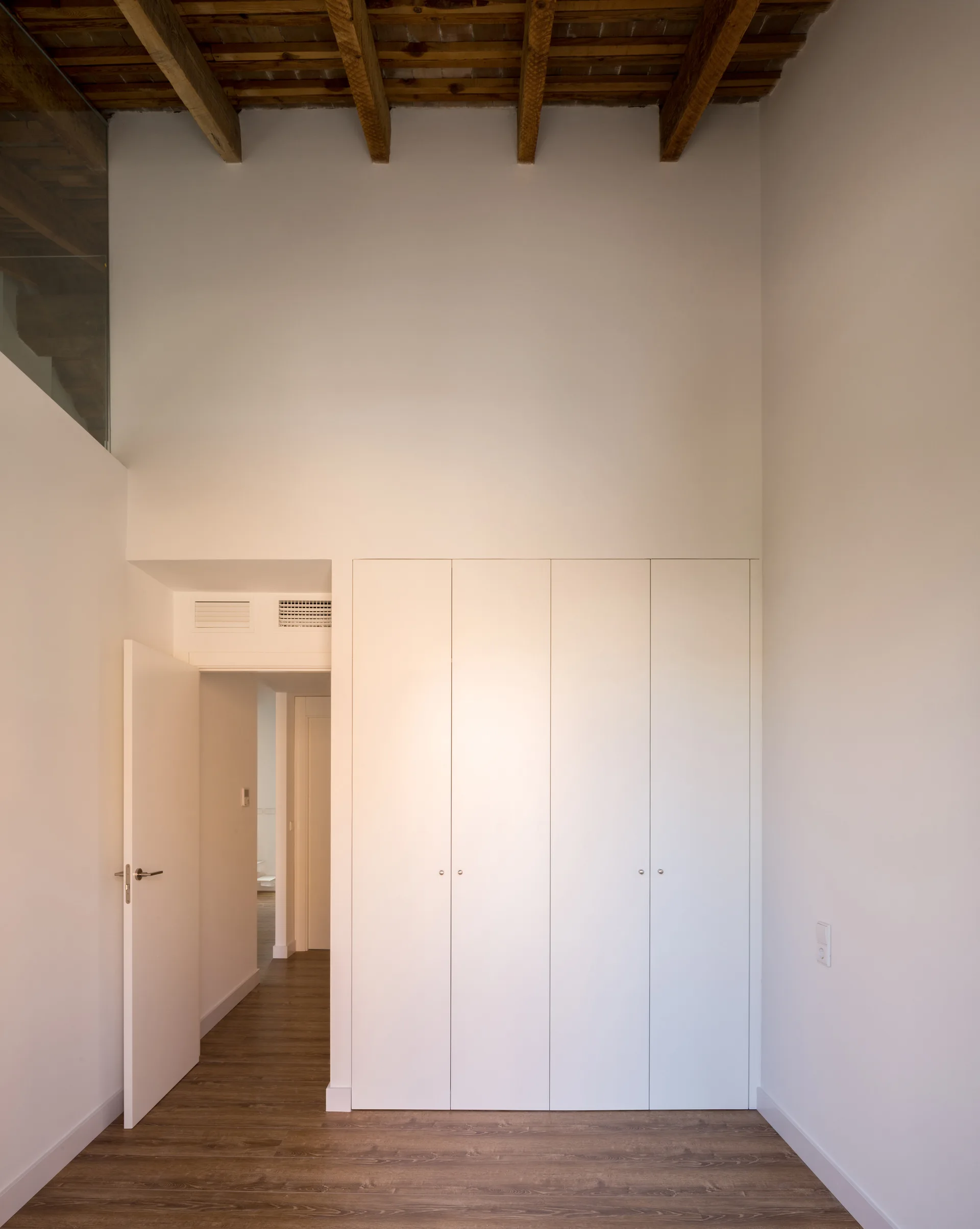 A minimalist white hallway featuring floor-to-ceiling built-in cabinets, an open doorway, dark wood floors, and exposed wooden ceiling beams.