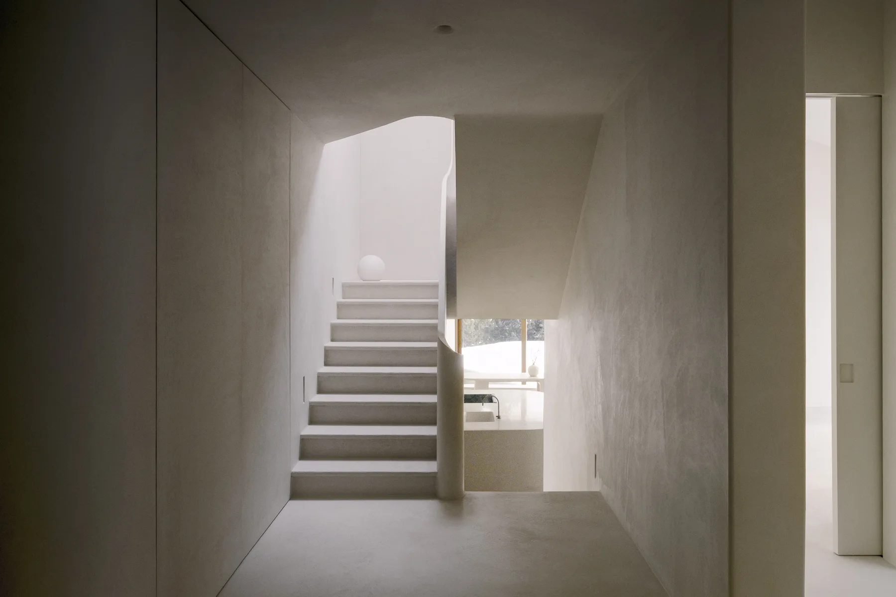 A minimalist off-white plaster staircase features a seamless sculptural design and a spherical lamp on an upper landing.