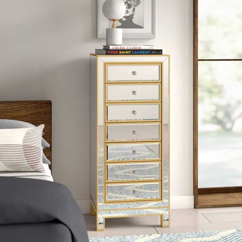 A tall mirrored chest of drawers with gold metal trim sits in a bedroom corner beside a wooden headboard and a patterned rug.