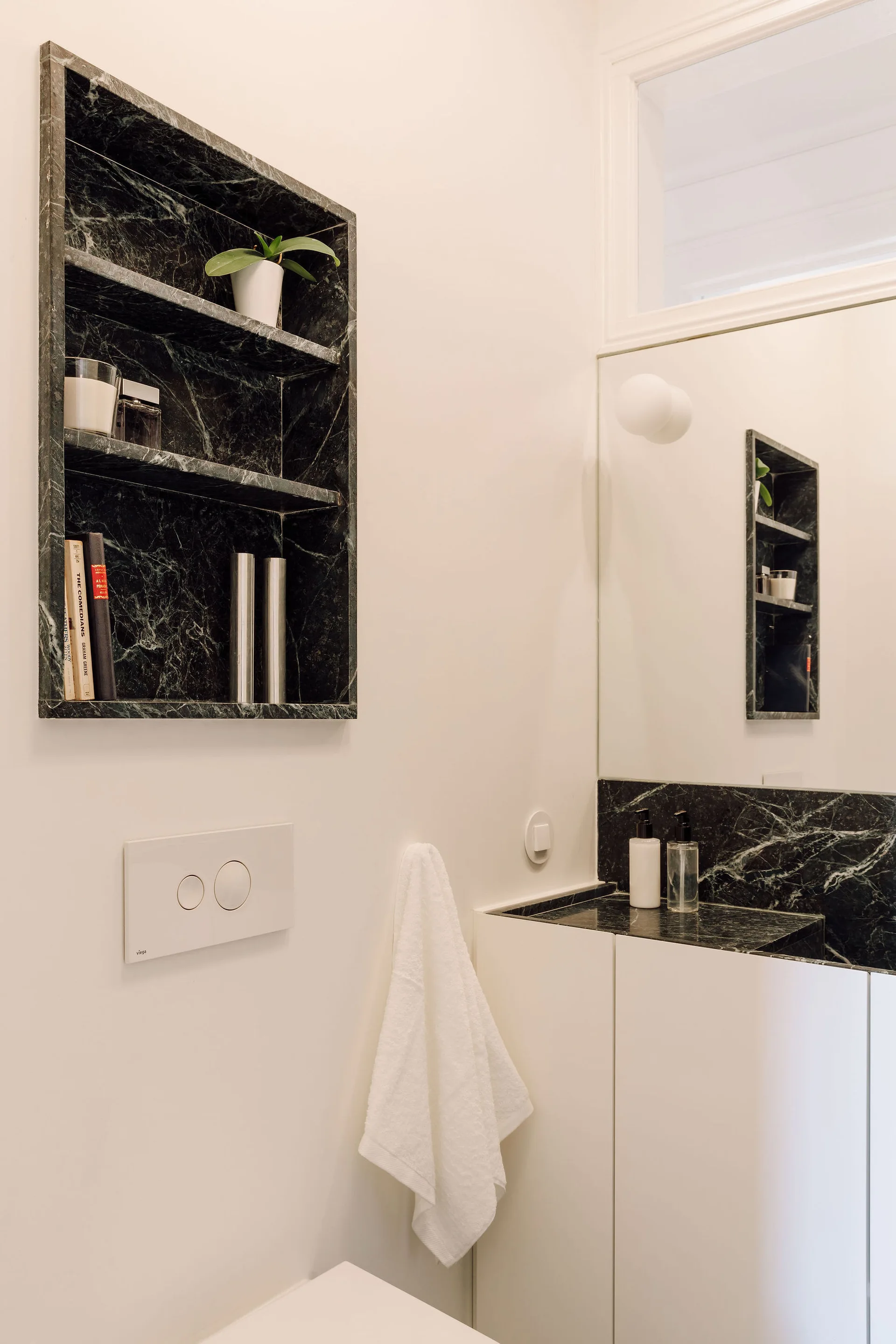 A minimalist powder room features a white wall with a recessed dark green marble shelf, a matching marble vanity, and a large mirror.