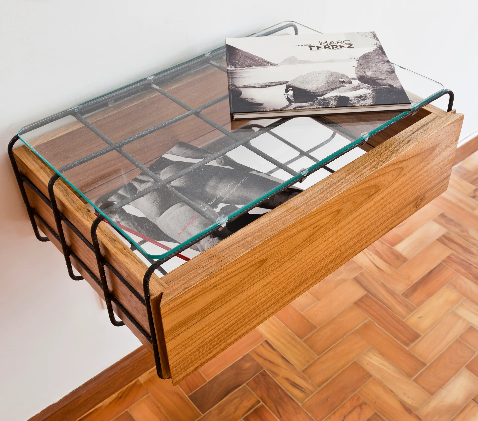 A wall-mounted wooden drawer featuring a black rebar frame and clear glass top is positioned against a white wall above a herringbone wood floor.