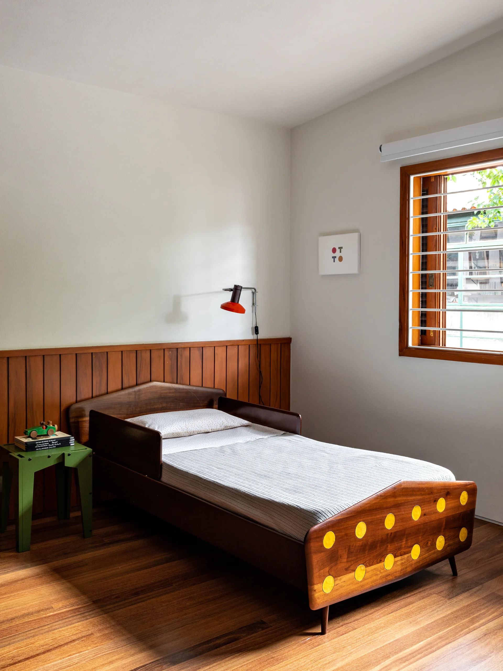 A child's wooden bed with yellow circle details sits against a wood-paneled wall next to a green metal side table and a red wall sconce.