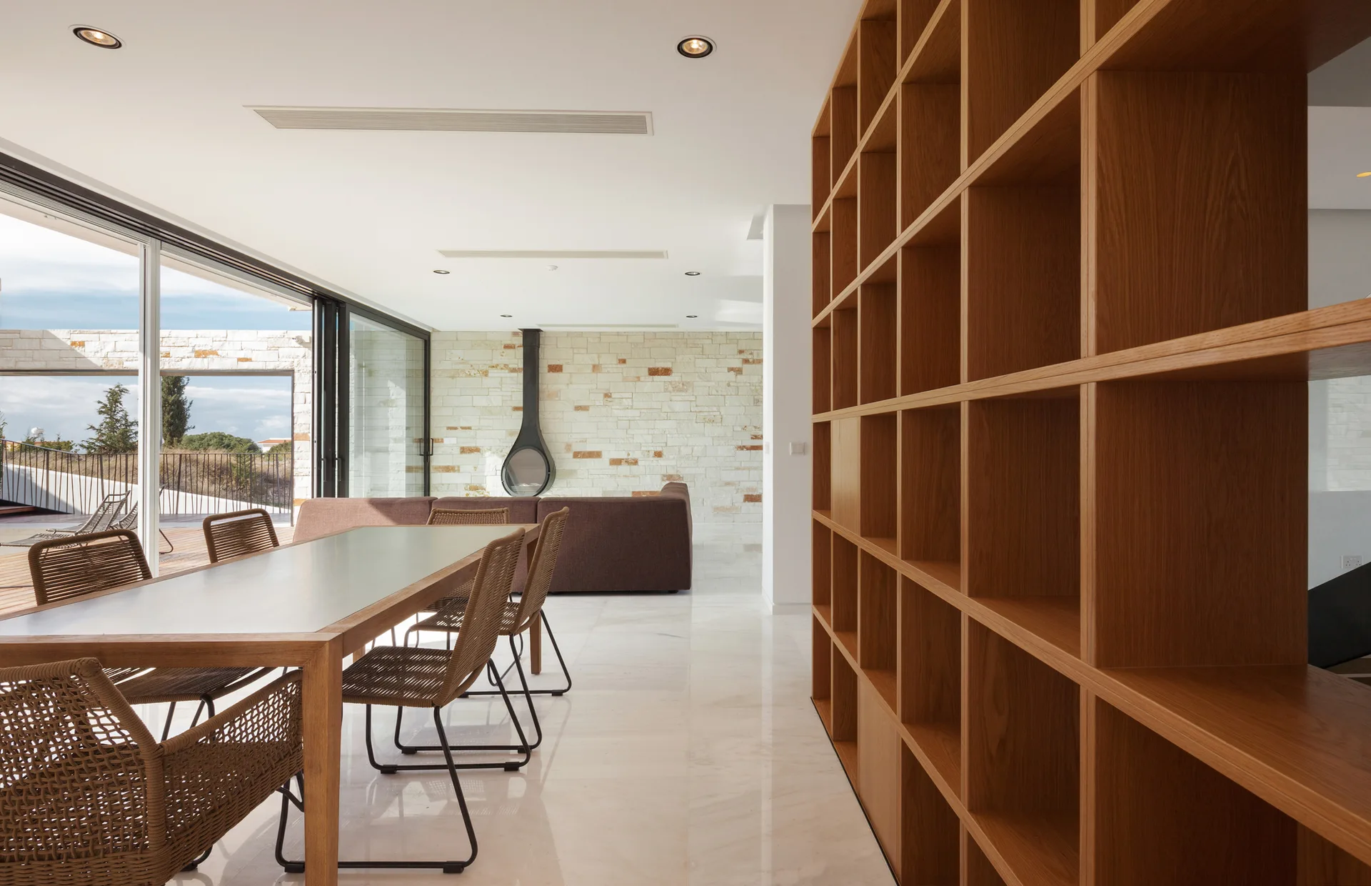 A modern dining room featuring a wooden table with woven chairs, a floor-to-ceiling grid bookshelf, and a fireplace in the background.