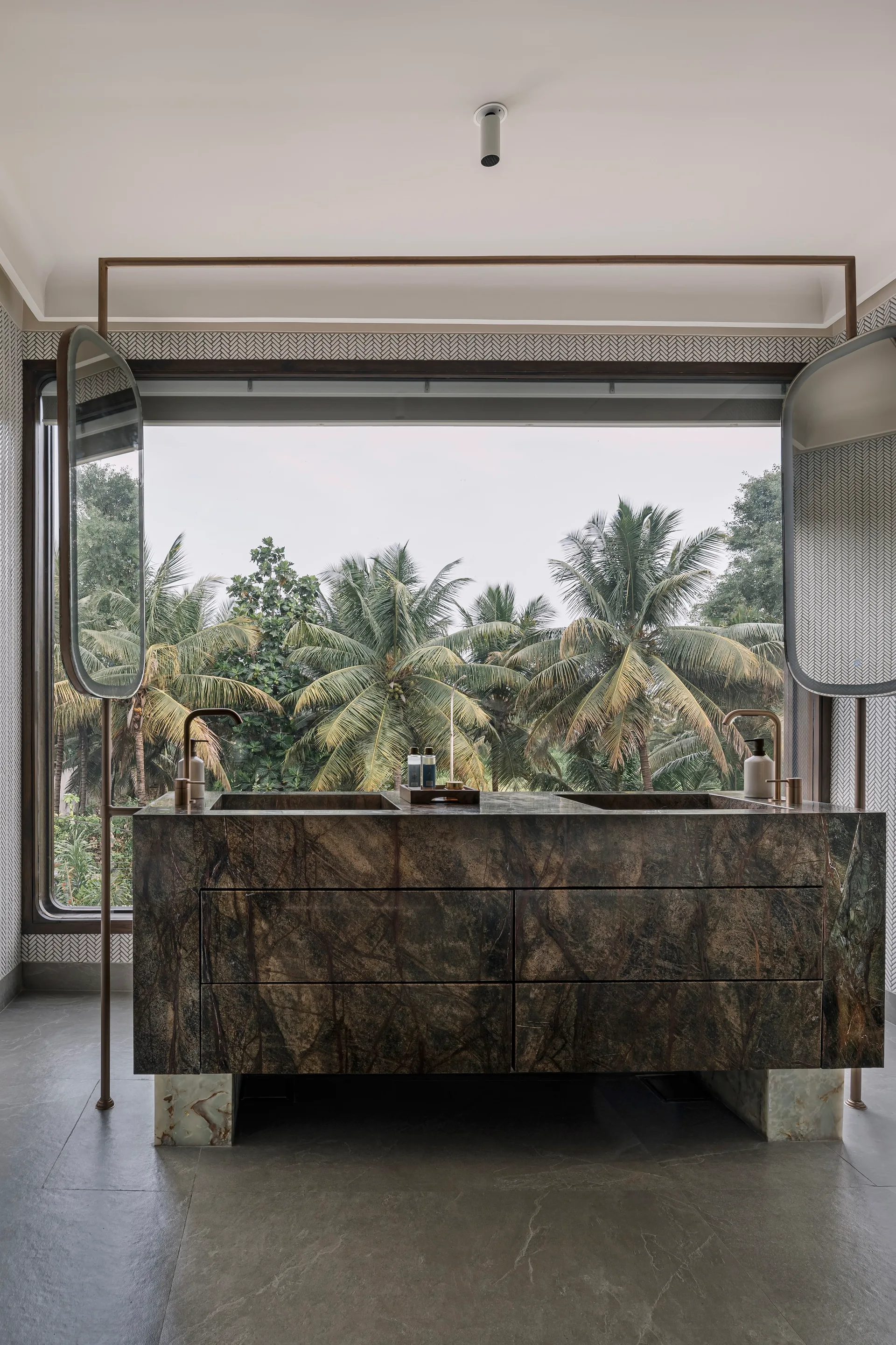 A modern bathroom features a dark veined stone double-sink vanity set against a large window framing a view of lush green palm trees.
