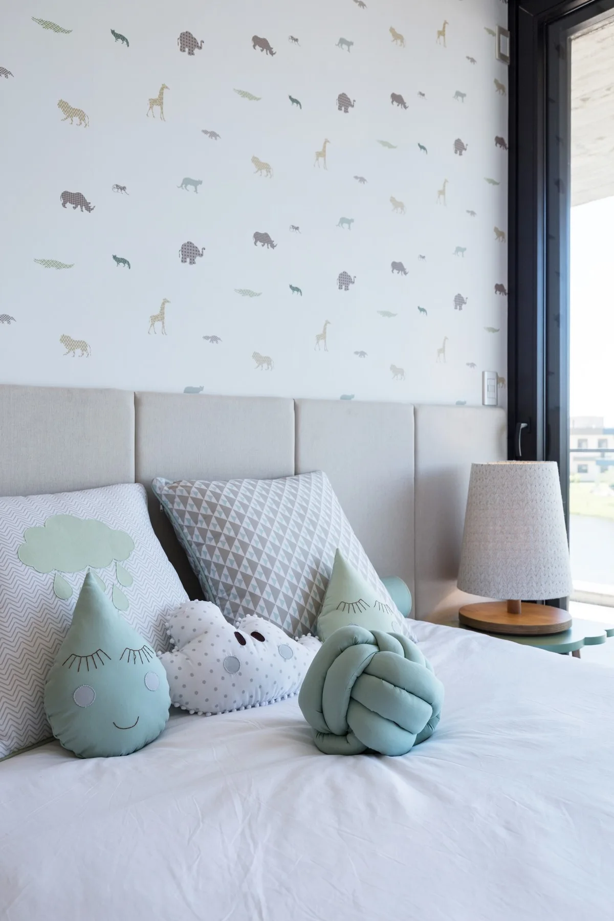 A child's bed featuring a light beige paneled headboard, animal print wallpaper, and several whimsical pastel green throw pillows.