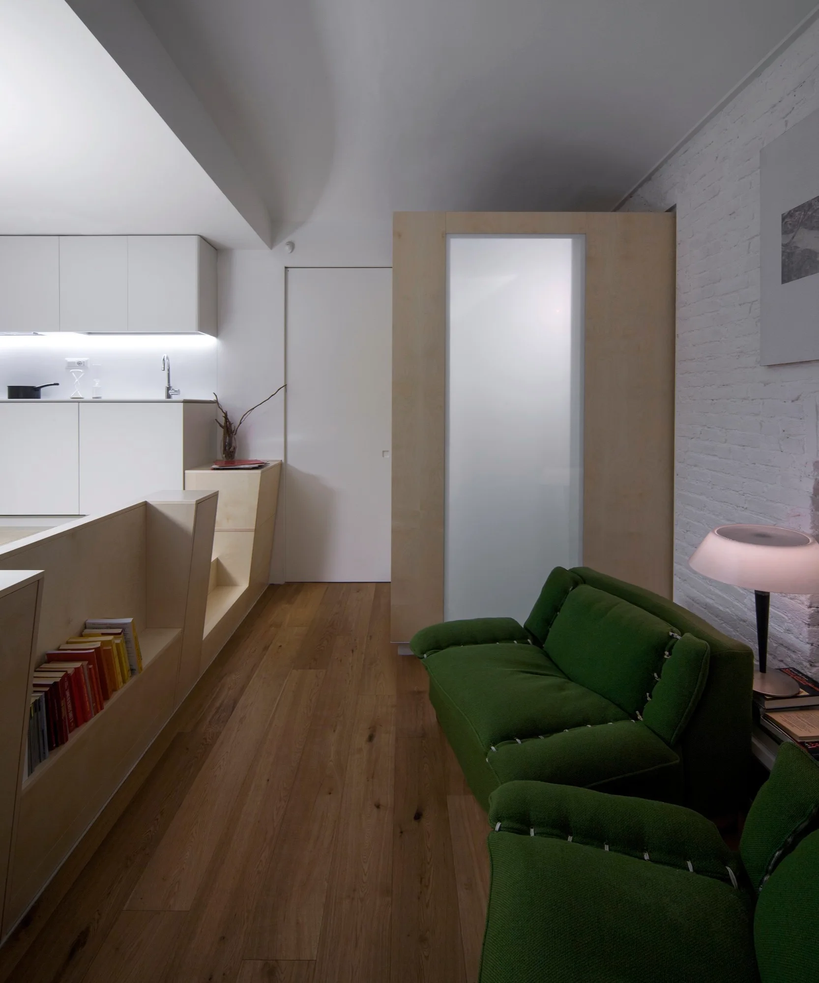 A modern living area featuring green armchairs, a white painted brick wall with a lamp, and custom plywood partition walls near a kitchen.