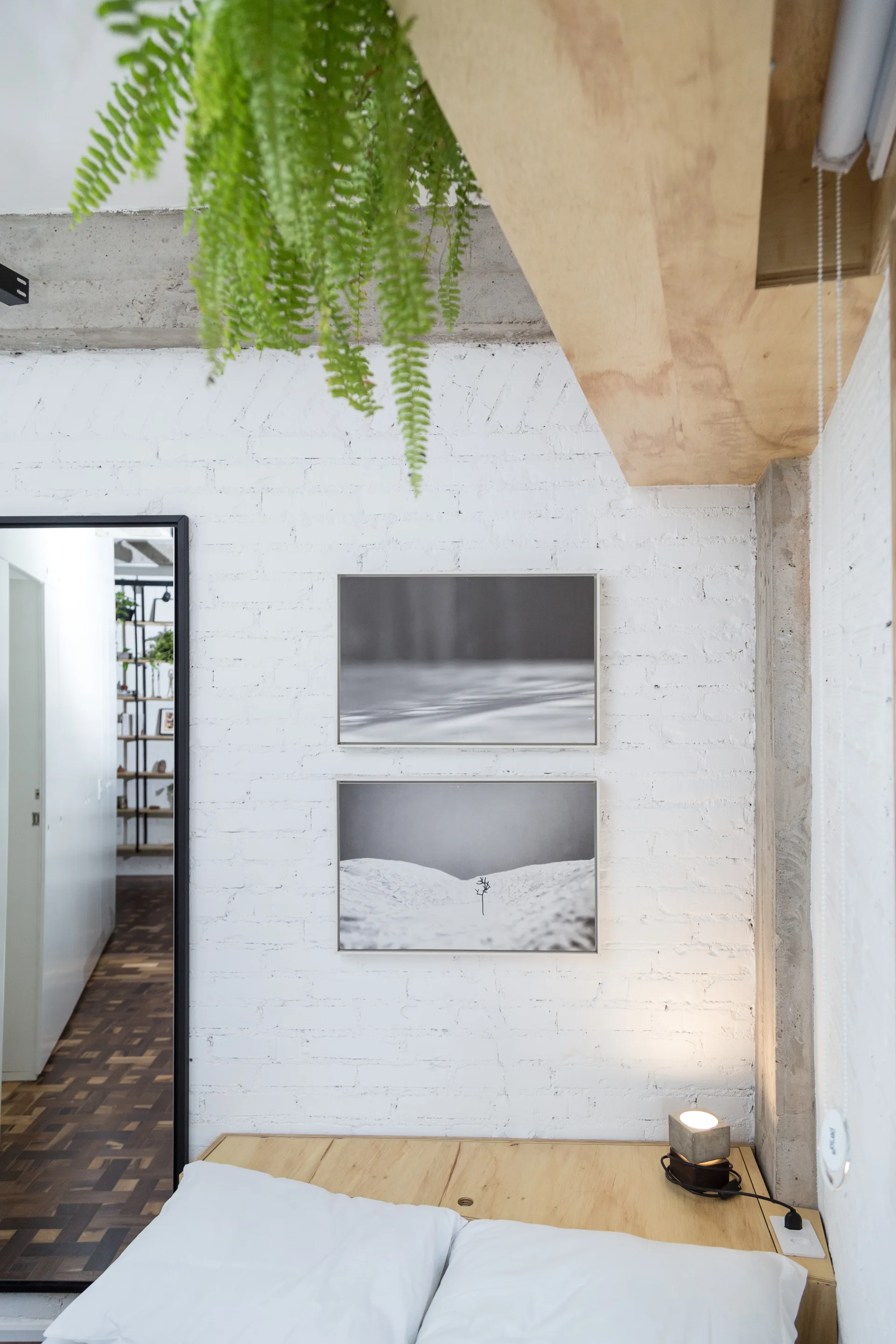 A minimalist bedroom featuring a white painted brick wall decorated with two stacked black and white framed prints and a hanging fern.