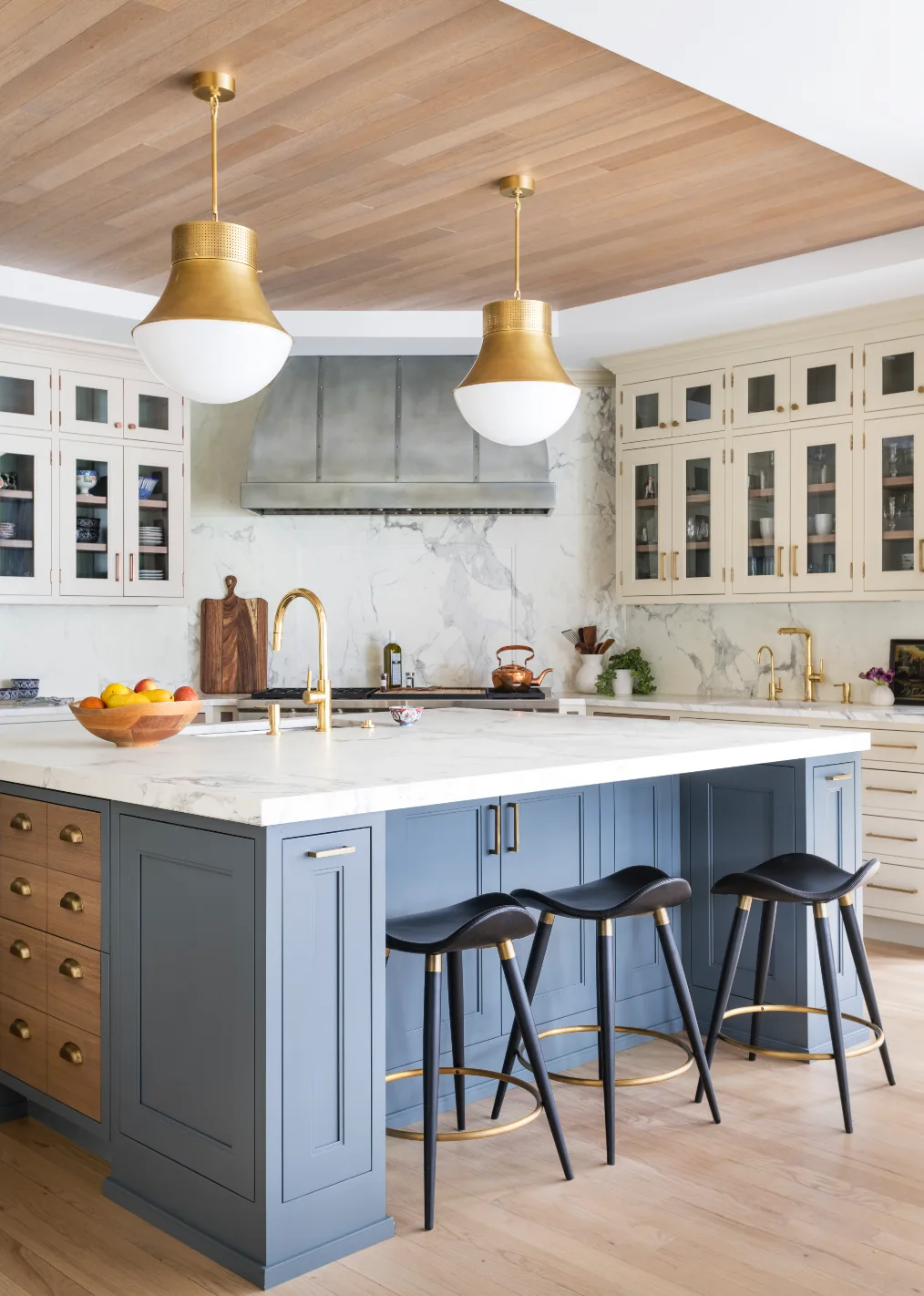 A modern transitional kitchen features a blue island with white marble countertops, black bar stools, brass pendant lights, and a wood plank ceiling.