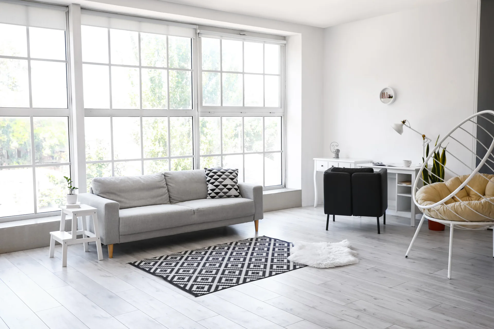 A bright Scandinavian-style living room features a grey sofa, a geometric patterned rug, and a white hanging chair beside a desk under large grid windows.