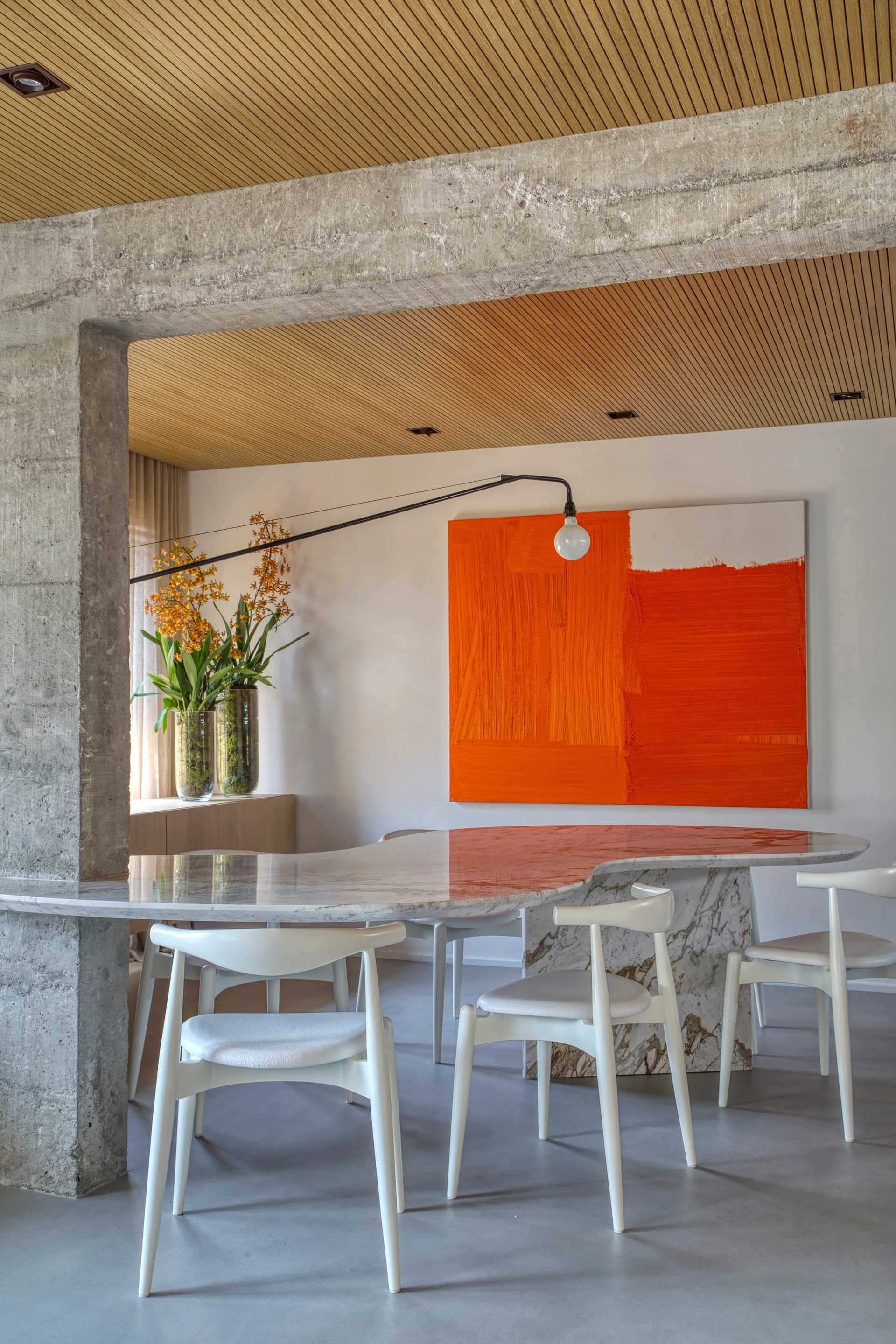A minimalist dining room features a curved marble table, white chairs, a large orange painting, and an exposed concrete beam under a slatted wood ceiling.