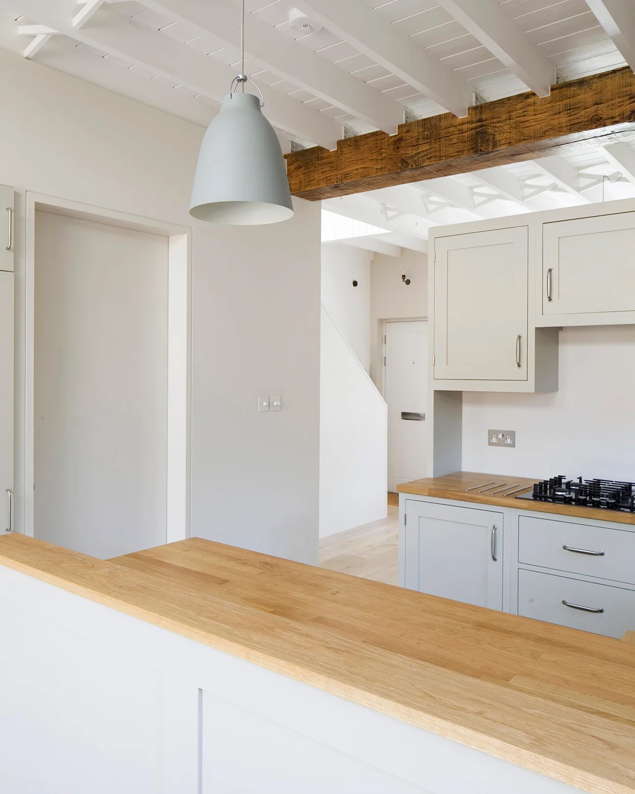 A modern farmhouse kitchen featuring light grey cabinetry, natural wood countertops, and a rustic exposed ceiling beam against painted white joists.