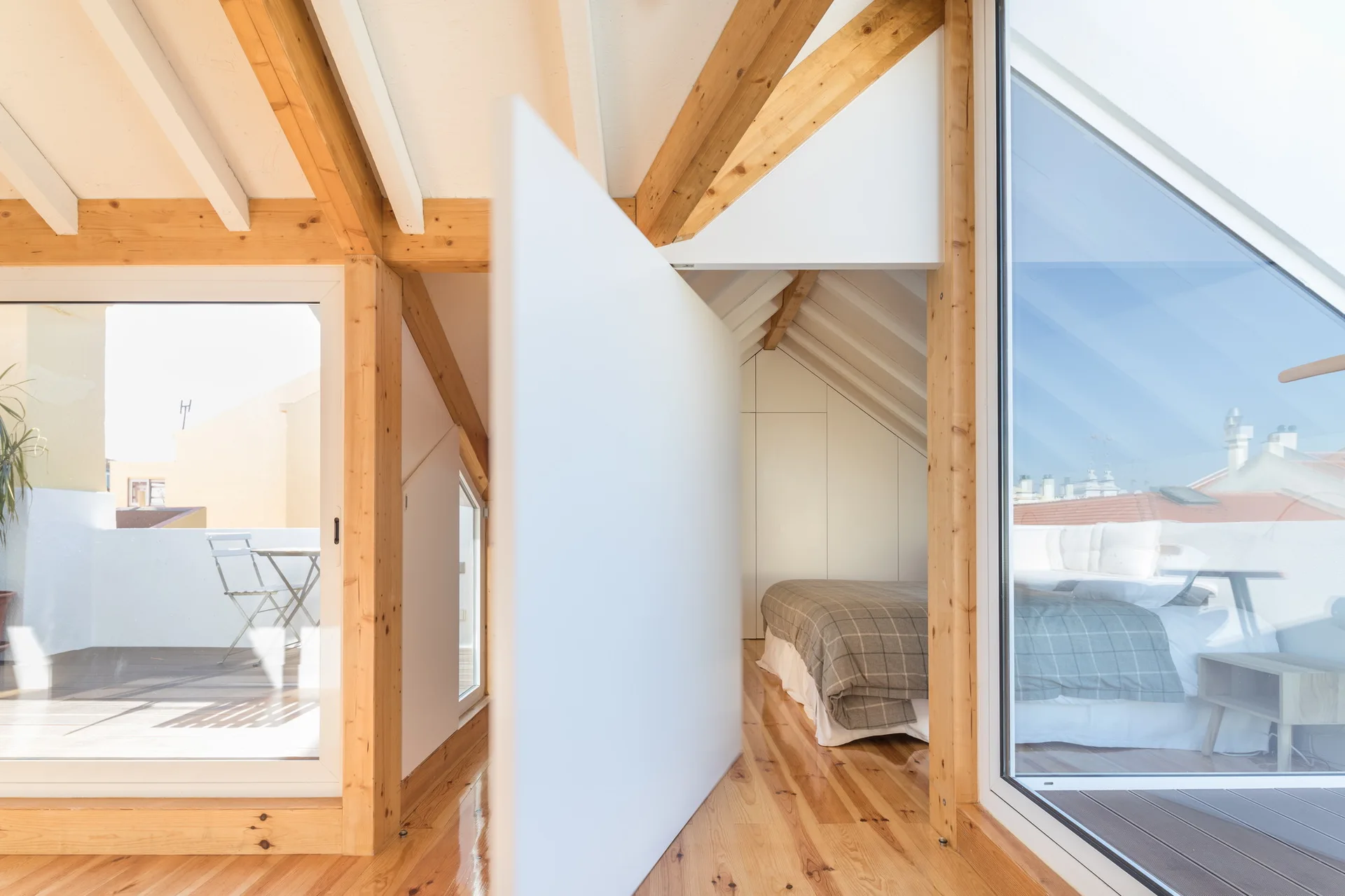 A bright attic bedroom with exposed timber beams, a bed with plaid bedding, glossy wood floors, and large glass doors leading to a balcony.