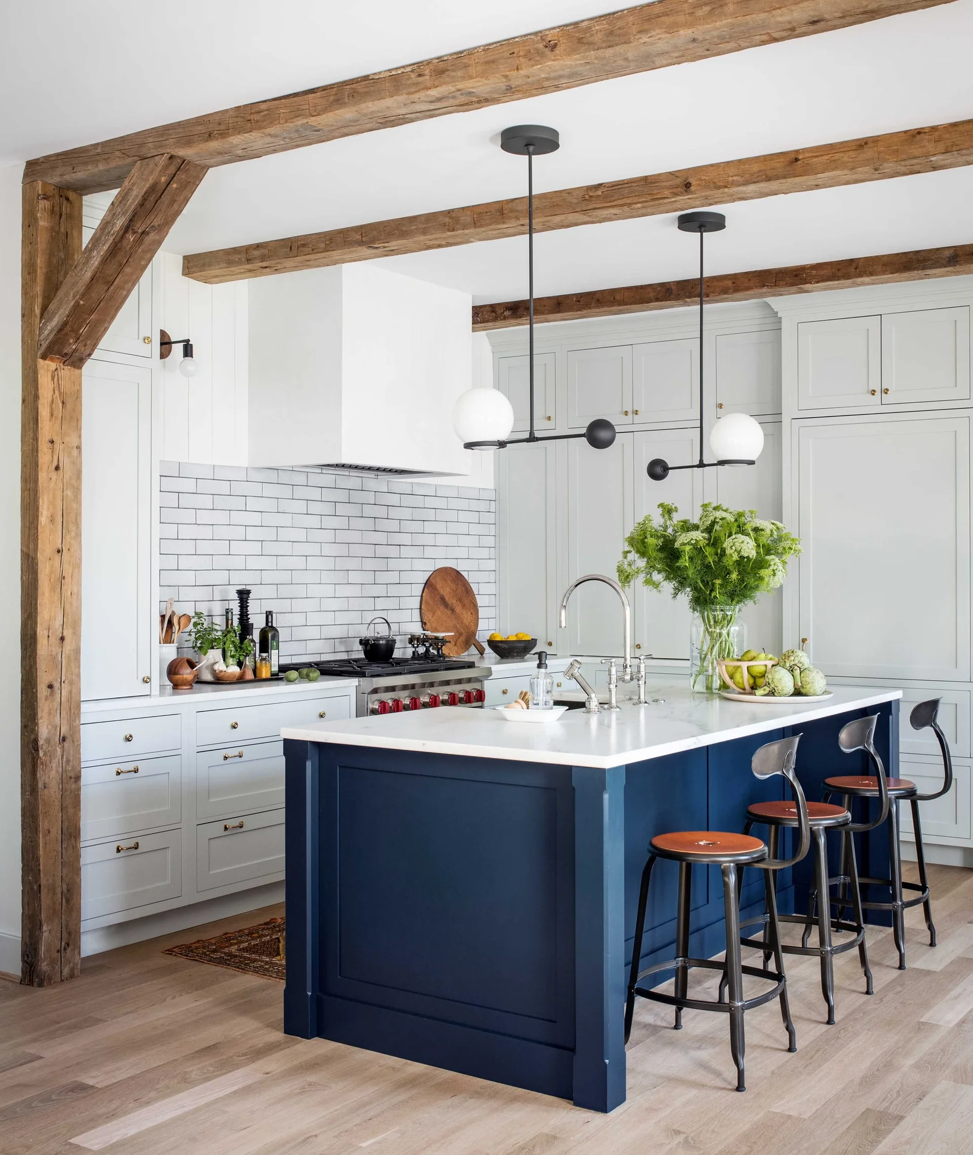 A light gray kitchen features a navy blue island with bar stools beneath rustic wood ceiling beams and modern black pendant lights.