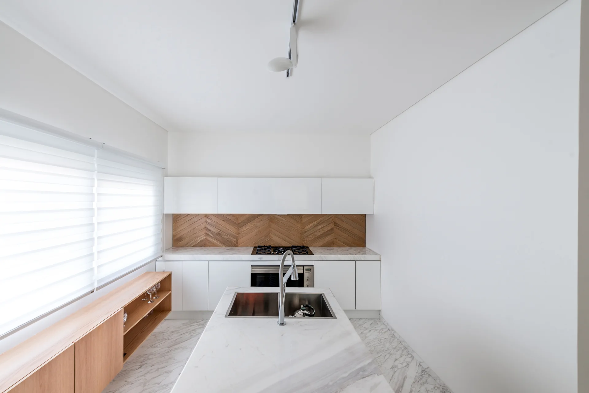 A modern white kitchen featuring a marble-topped island, flat-front cabinets, and a warm herringbone wood backsplash.