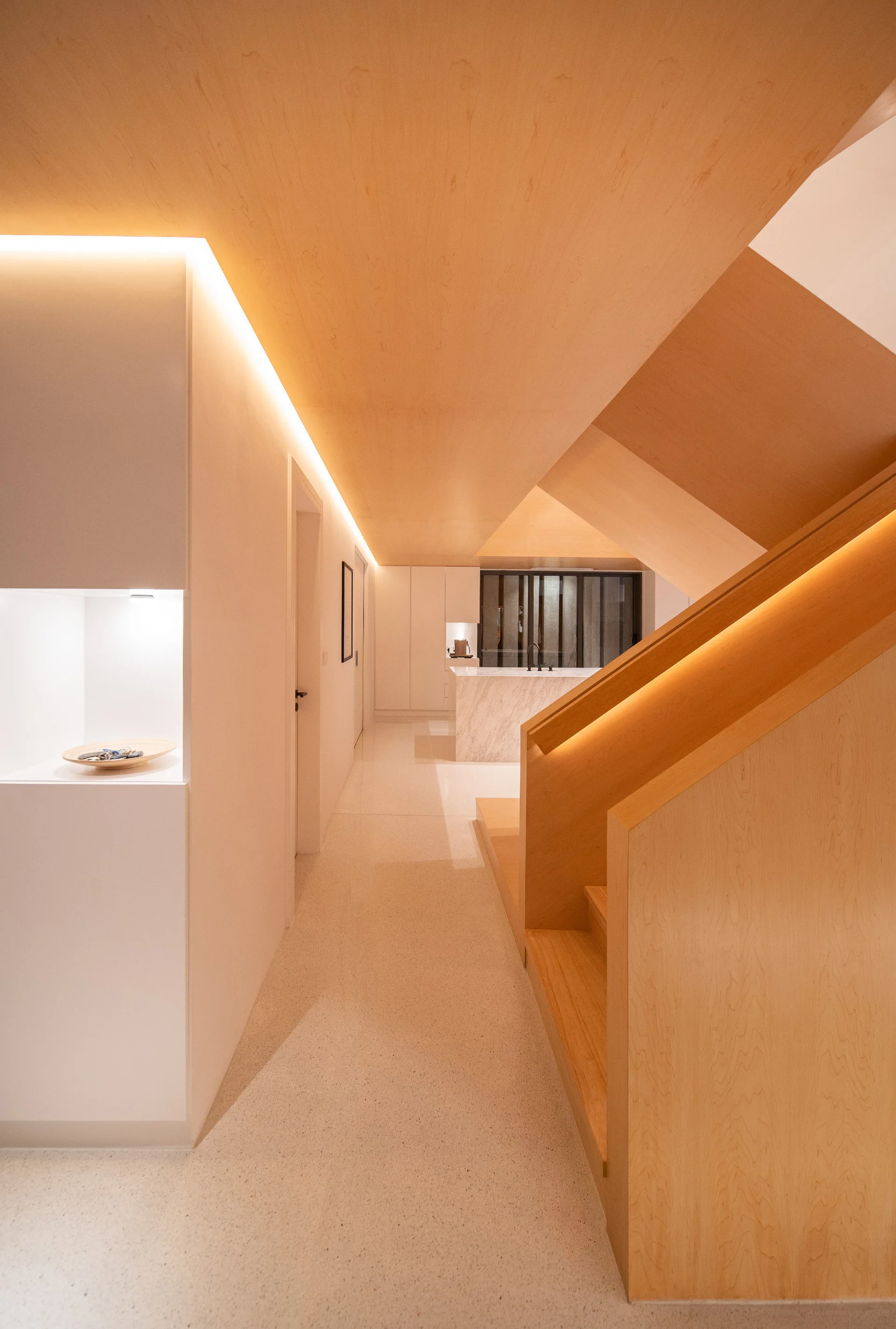 A modern hallway features a light wood staircase and matching wood-clad ceiling, integrated LED strip lighting, and a recessed display niche on a white wall.