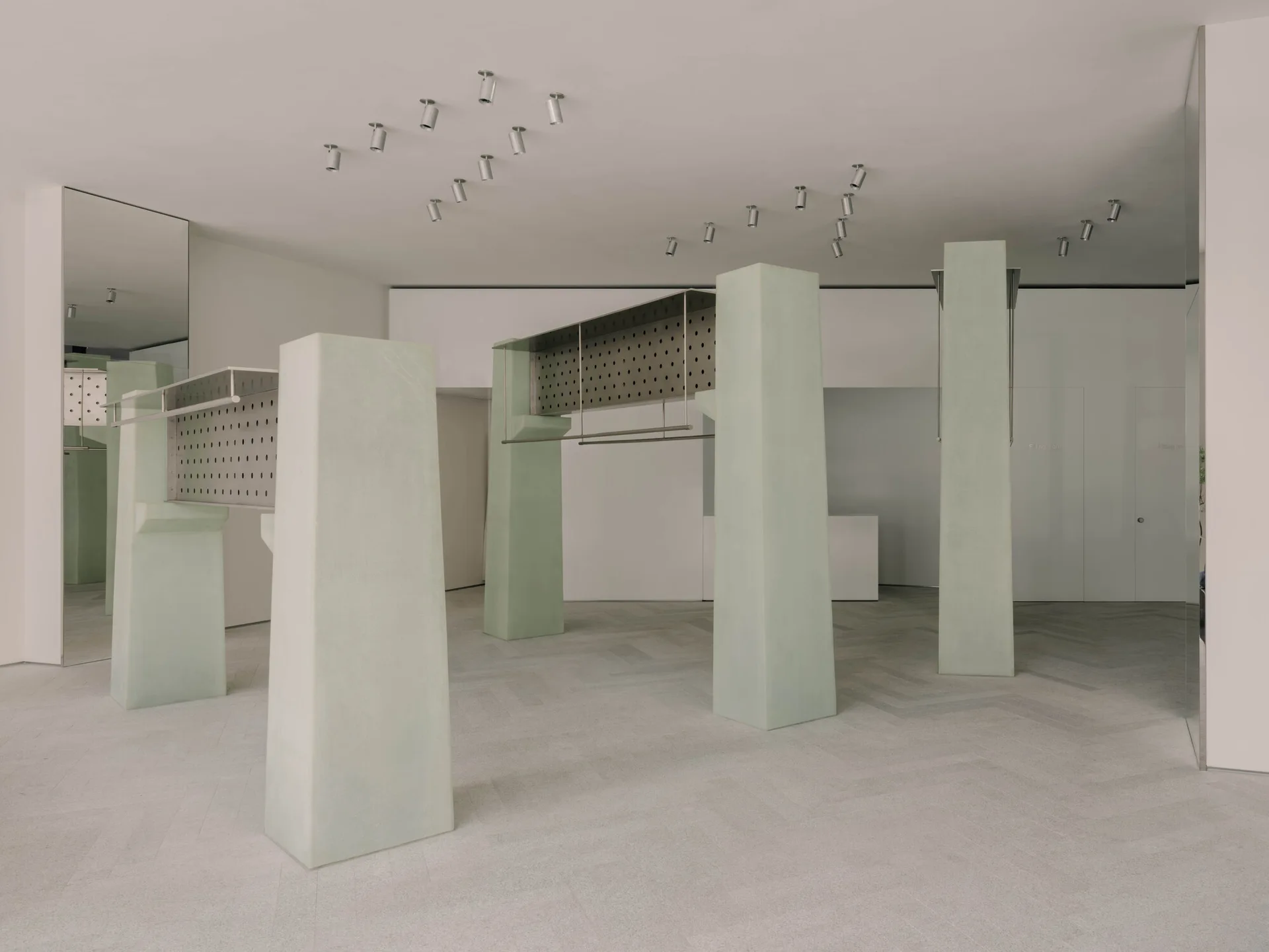 A minimalist retail showroom featuring pale green architectural pillars, suspended perforated metal racks, and light grey herringbone floor tiles.
