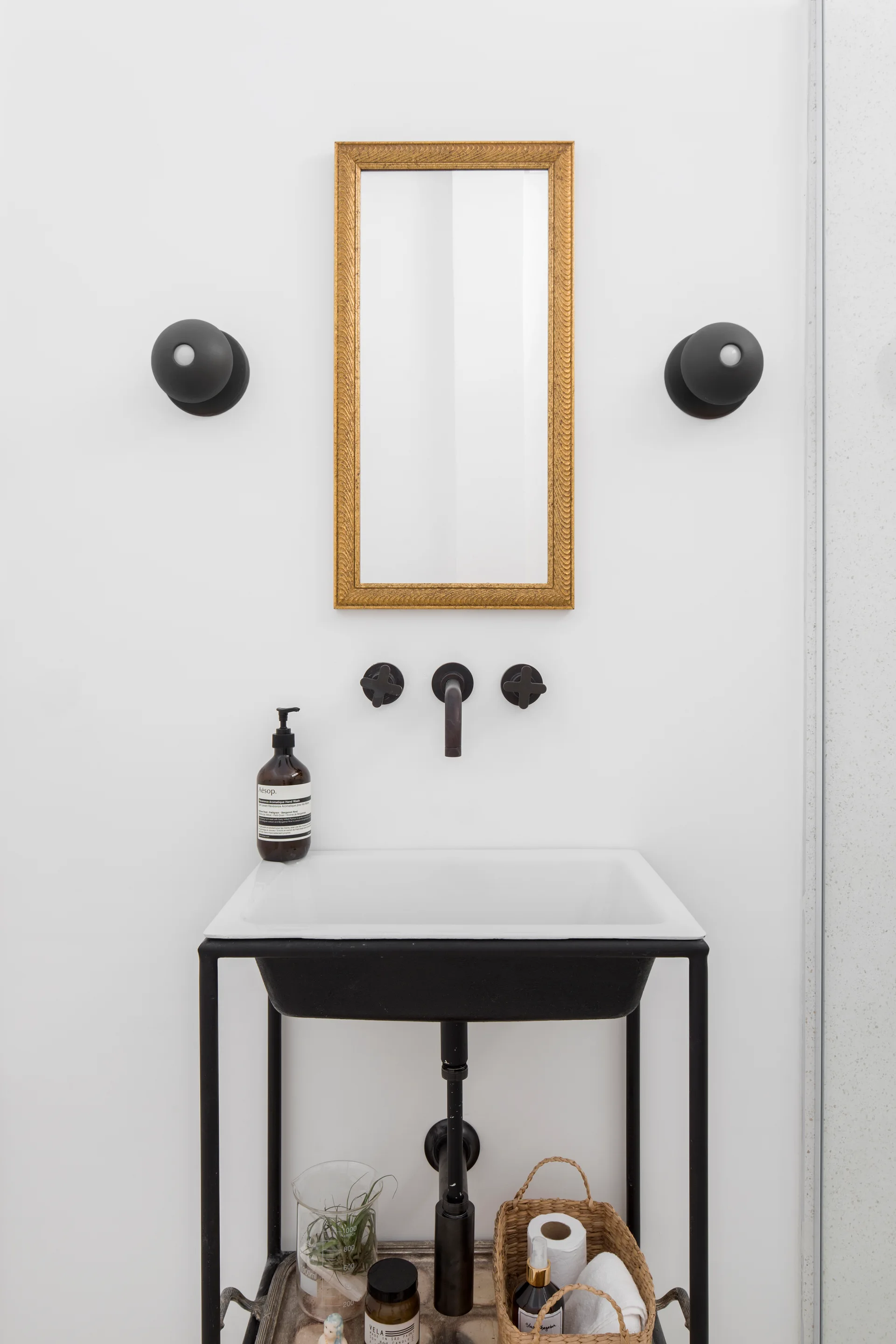 A minimalist white sink with a black metal console frame features a wall-mounted matte black faucet, flanked by spherical sconces beneath a gold-framed mirror.