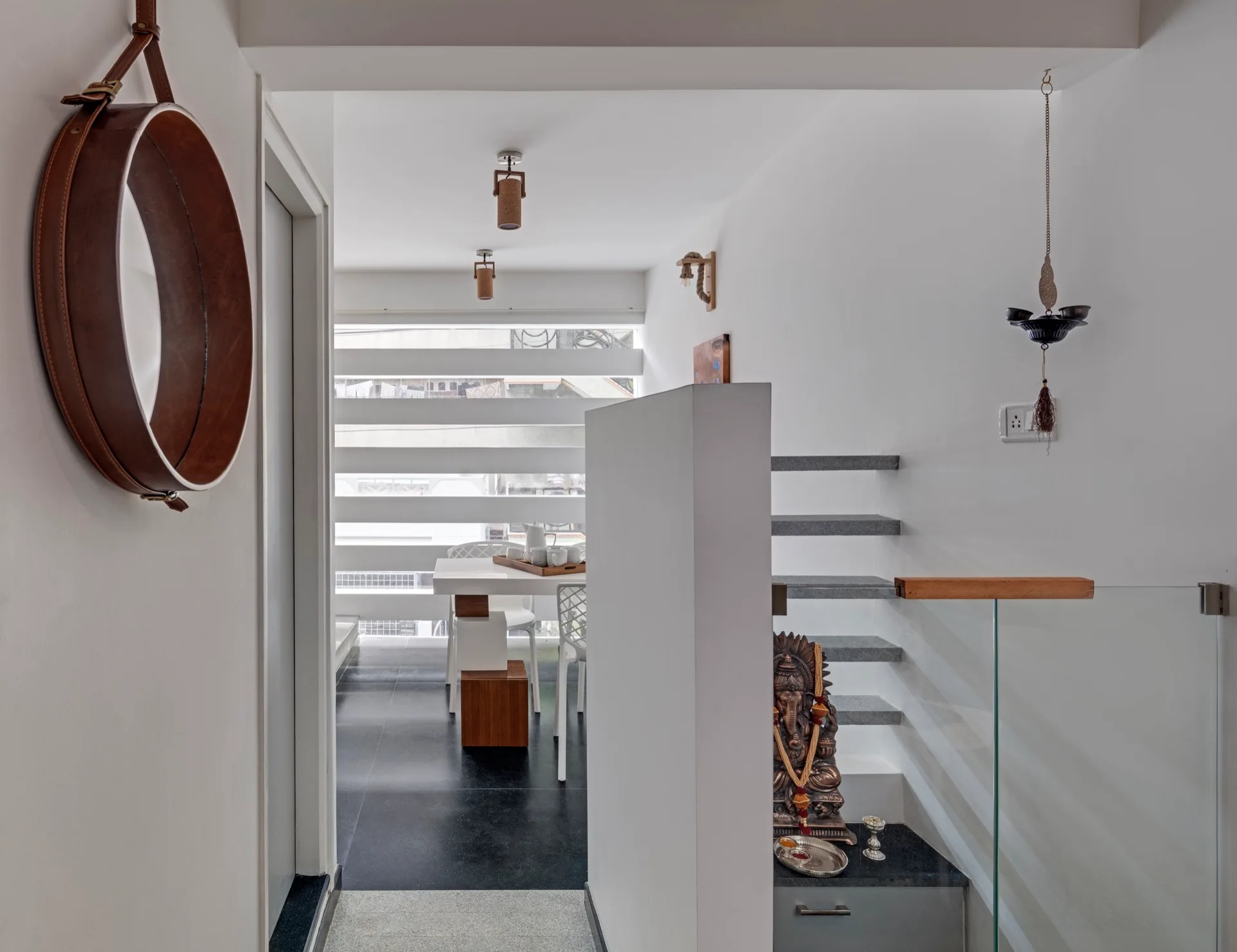 A modern white entryway features a round mirror hanging by a brown strap, opening to a dining area and a staircase with a glass railing.
