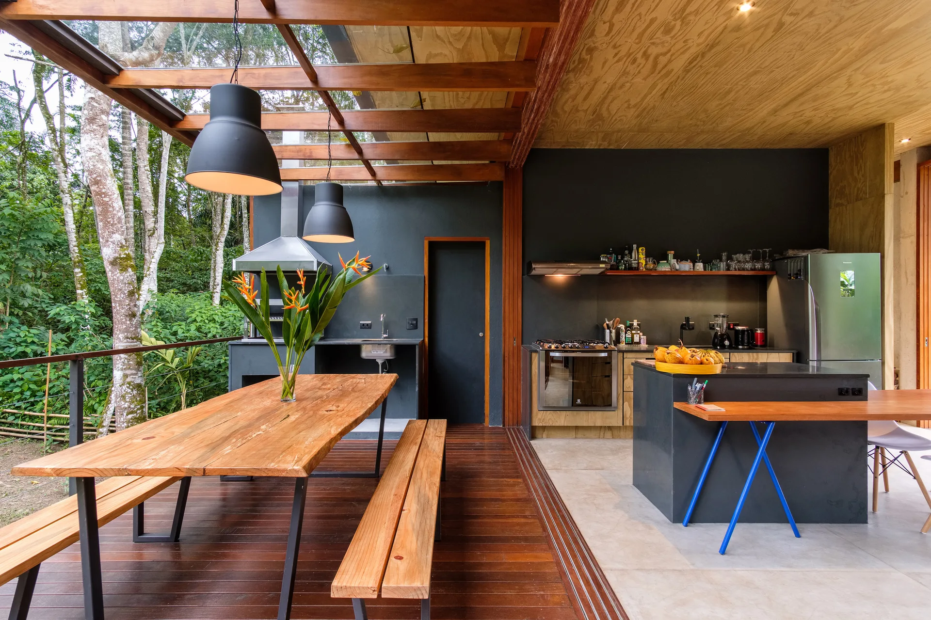 A covered outdoor dining deck with a long wood table and benches sits next to an indoor kitchen with dark charcoal walls and cabinetry.