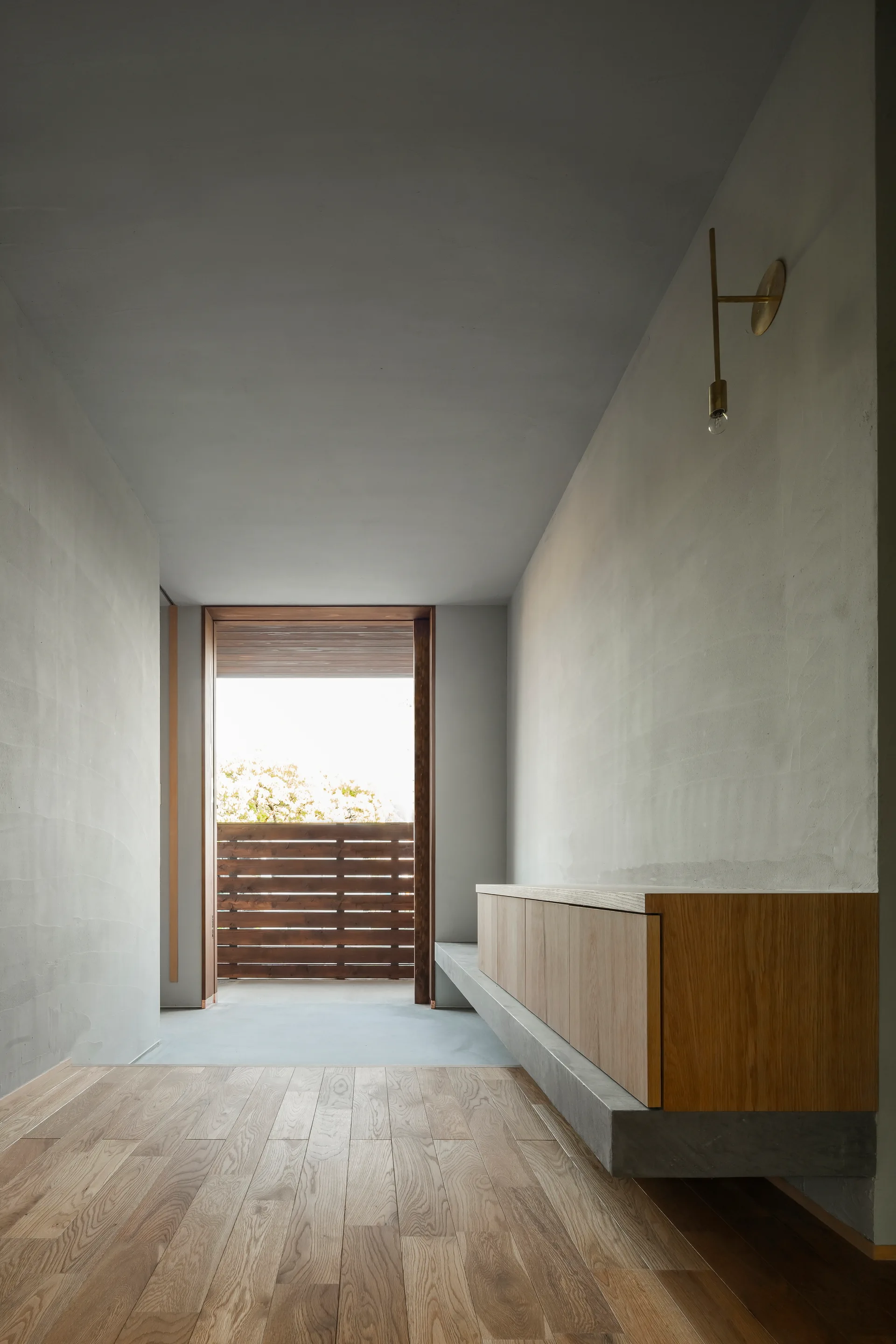 A minimalist hallway features light wood plank flooring, textured grey plaster walls, a floating wooden storage cabinet, and a view to a wood-slatted balcony.