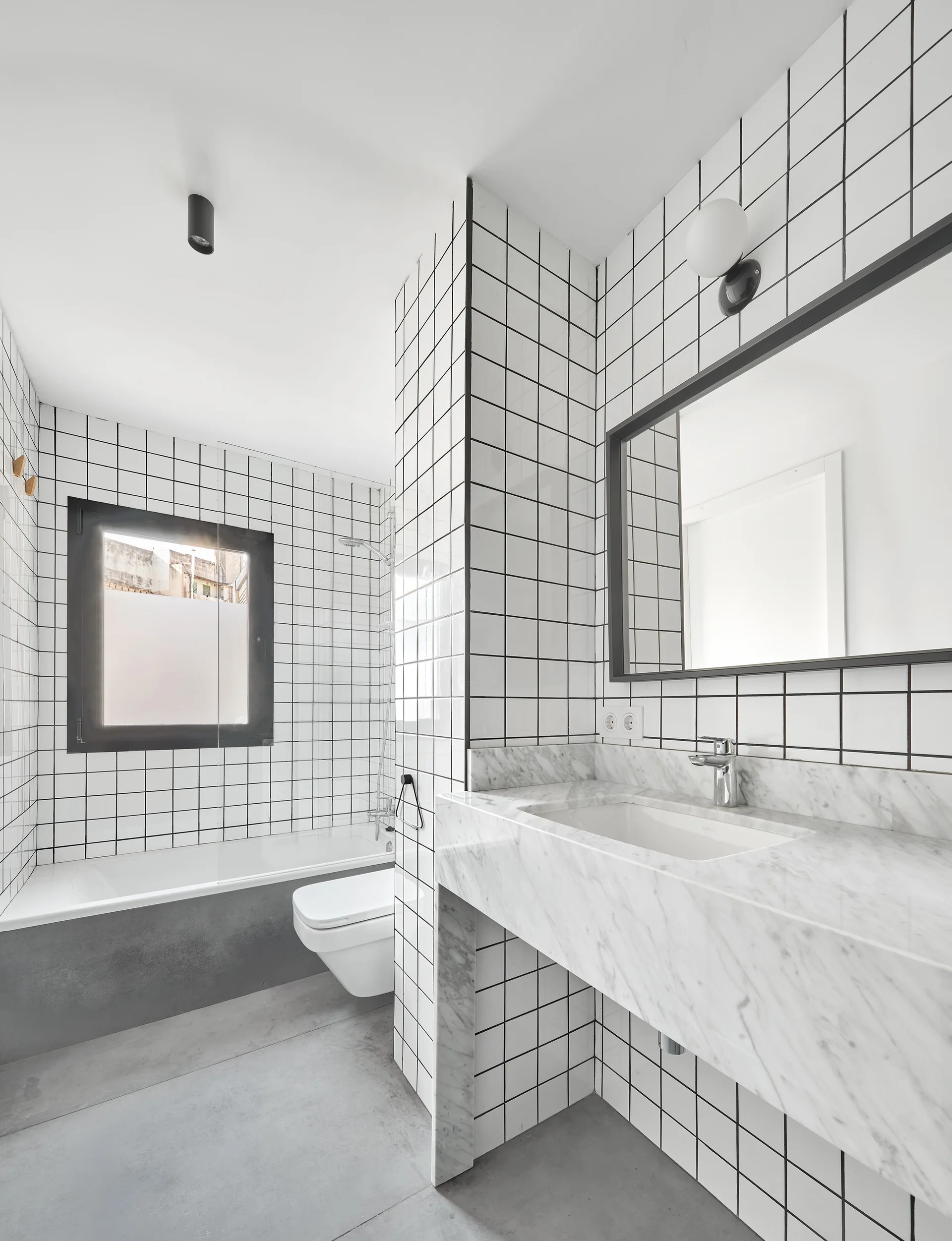 A minimalist bathroom features white grid wall tiles, a polished marble vanity, a wall-hung toilet, concrete floors, and a bathtub.
