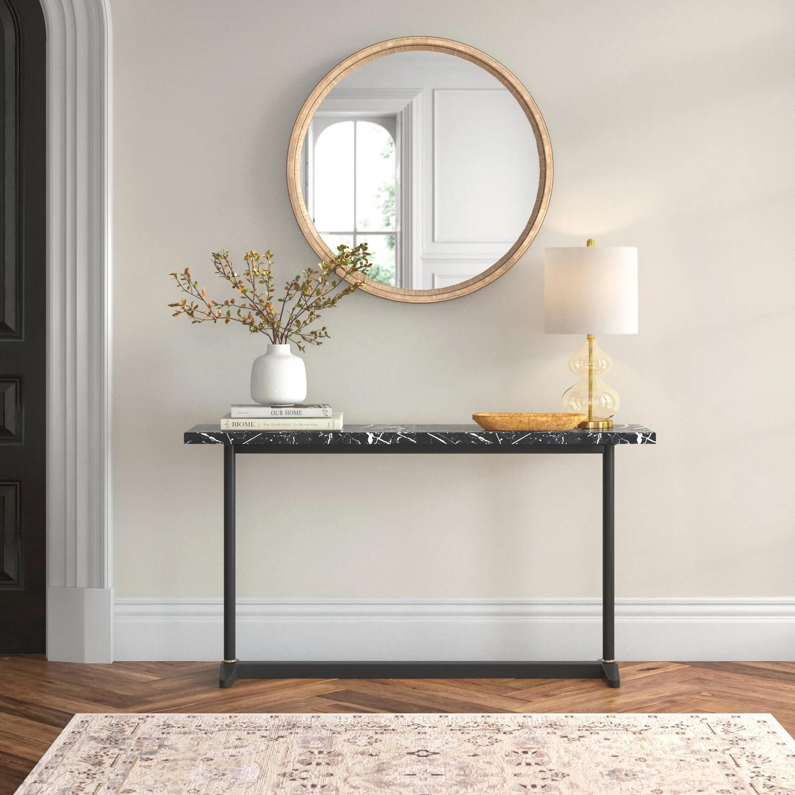 A black marble console table styled with a lamp, books, and vase stands beneath a round wood-framed mirror in a hallway with herringbone floors.