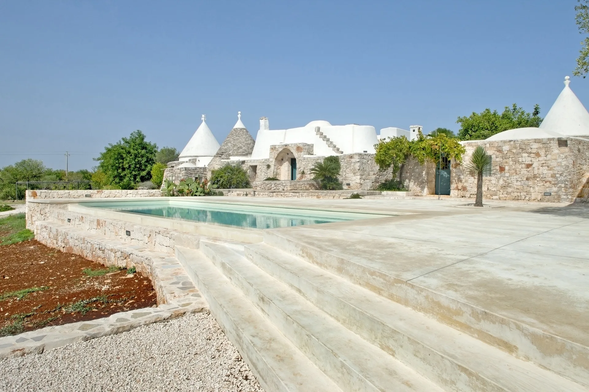 A rectangular swimming pool sits on a concrete terrace with wide steps, backed by stone walls and traditional white trulli houses under a blue sky.