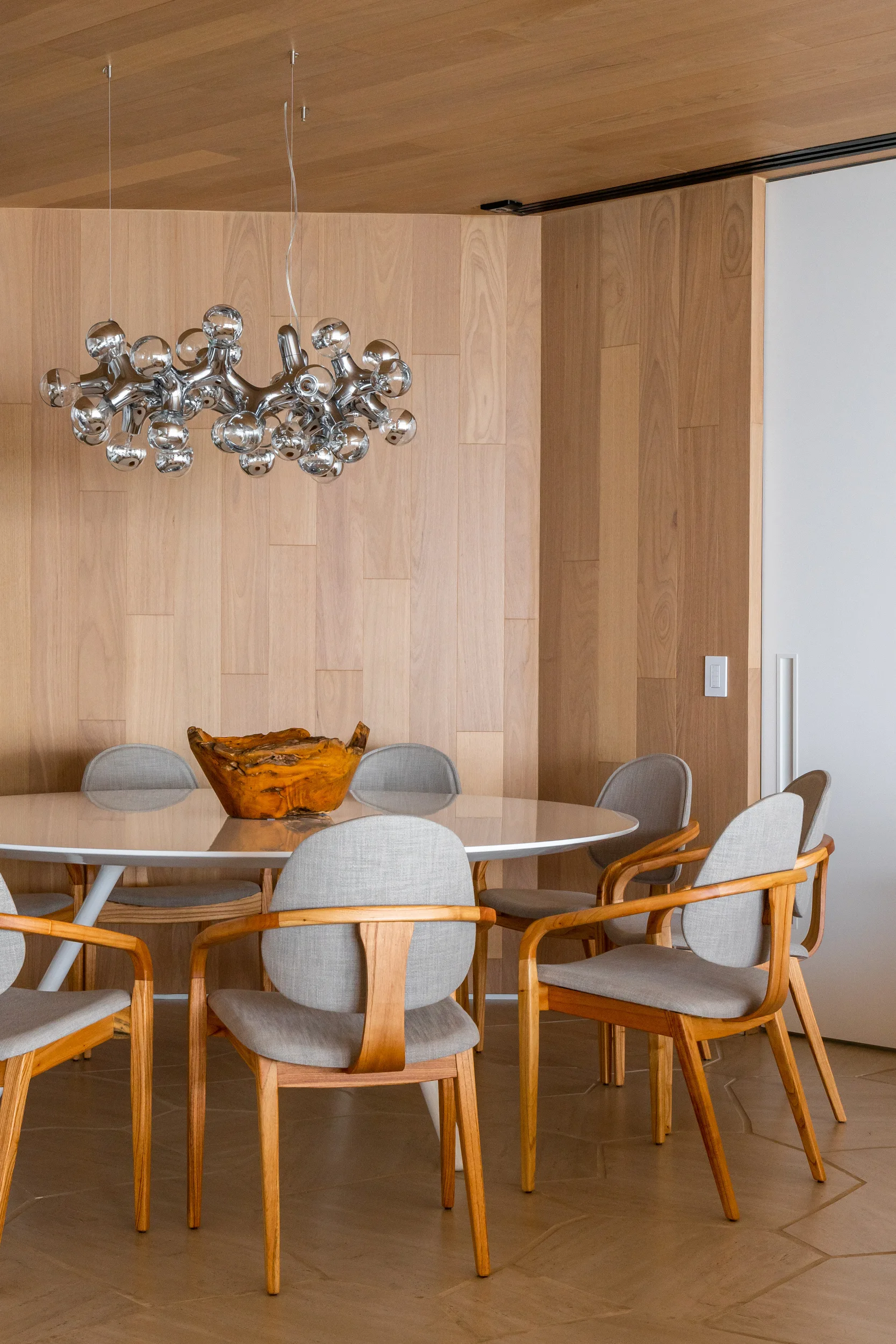 A modern dining room features a round white table, grey upholstered wooden chairs, a wood paneled wall, and a chrome molecular chandelier.