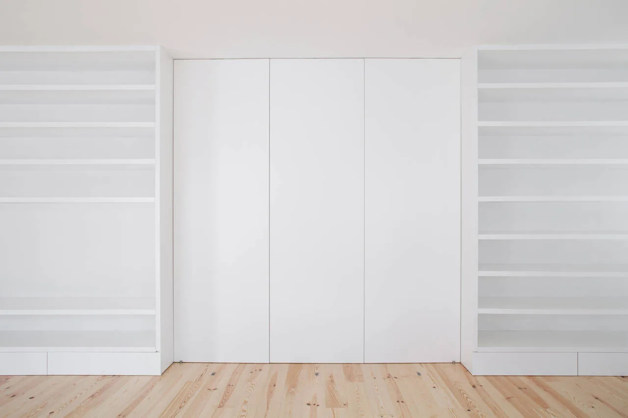 Minimalist white built-in shelving units flank seamless handleless closet doors on a light-colored natural wood plank floor.